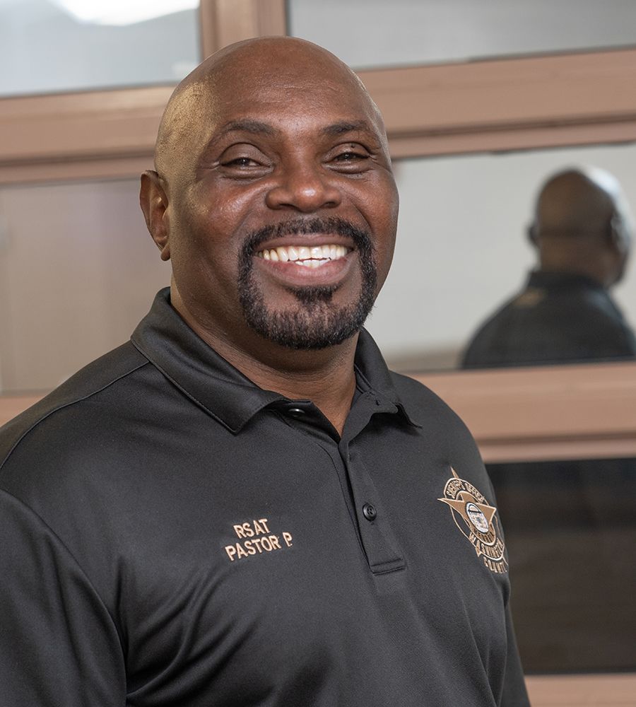 A smiling person wearing a black polo shirt embroidered with "Pastor P" and a logo. The setting includes a reflective surface in the background displaying the person's reflection.
