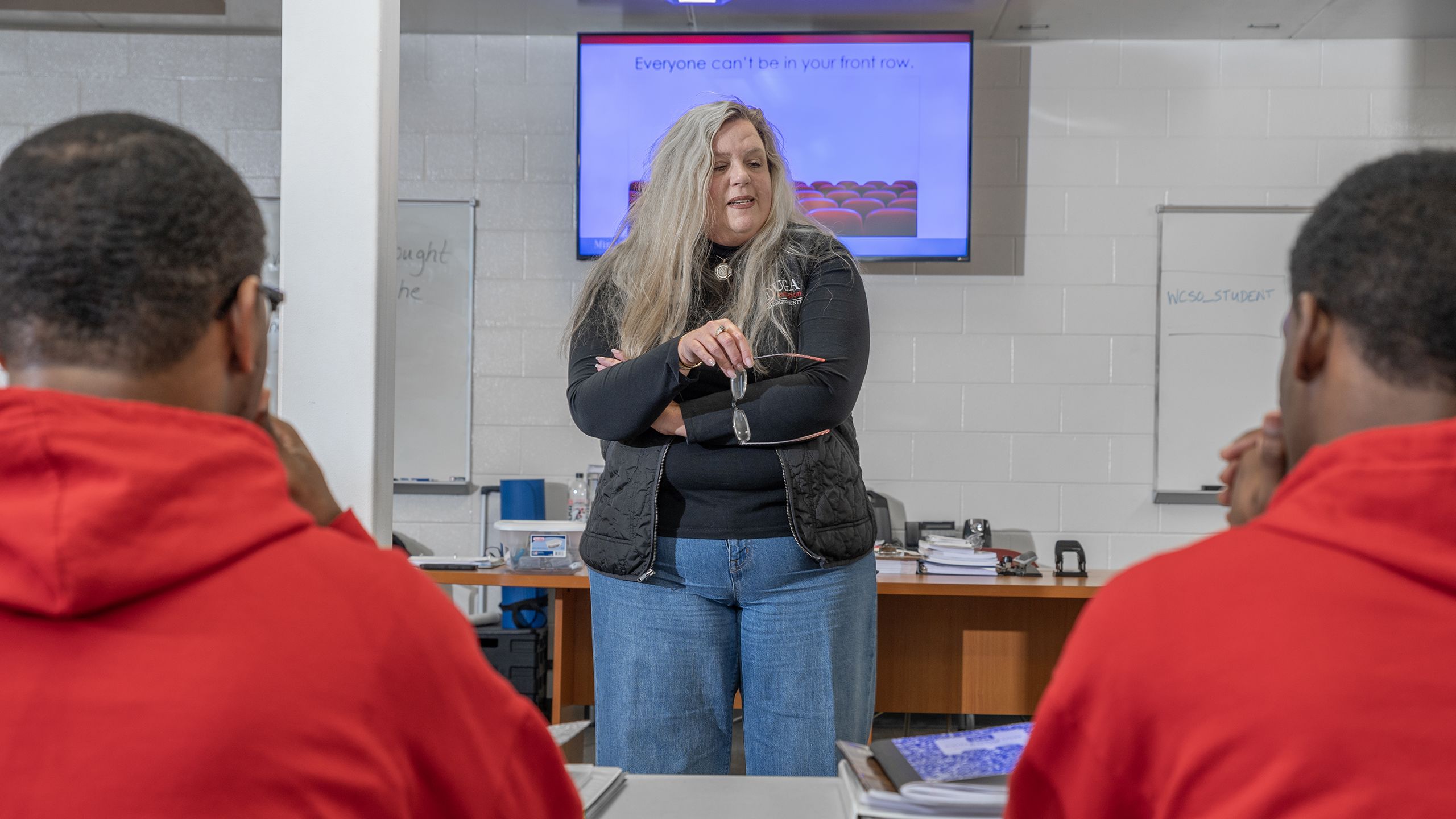 An instructor stands in front of a classroom, gesturing with a pen while students, wearing red uniforms, listen attentively; a PowerPoint presentation is visible on the screen in the background.