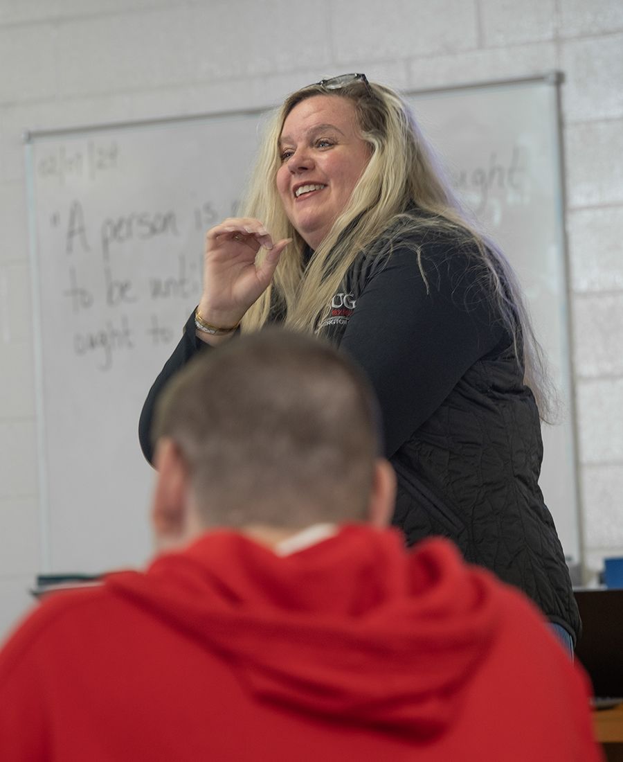 Teacher standing in front of a whiteboard, smiling and interacting with a student in a classroom. The whiteboard has text that reads: "A person is to be understood as a totality of thoughts."