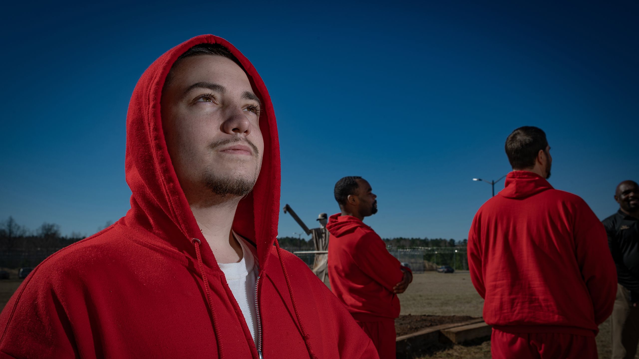 Three individuals outdoors during the day, all wearing red hoodies. The person in the foreground is looking upwards, lit by sunlight on the face, with a focused expression. A second person is visible in the mid-ground facing away, and a third person, partially visible, stands in the background also facing away. Clear blue sky in the background with no clouds.