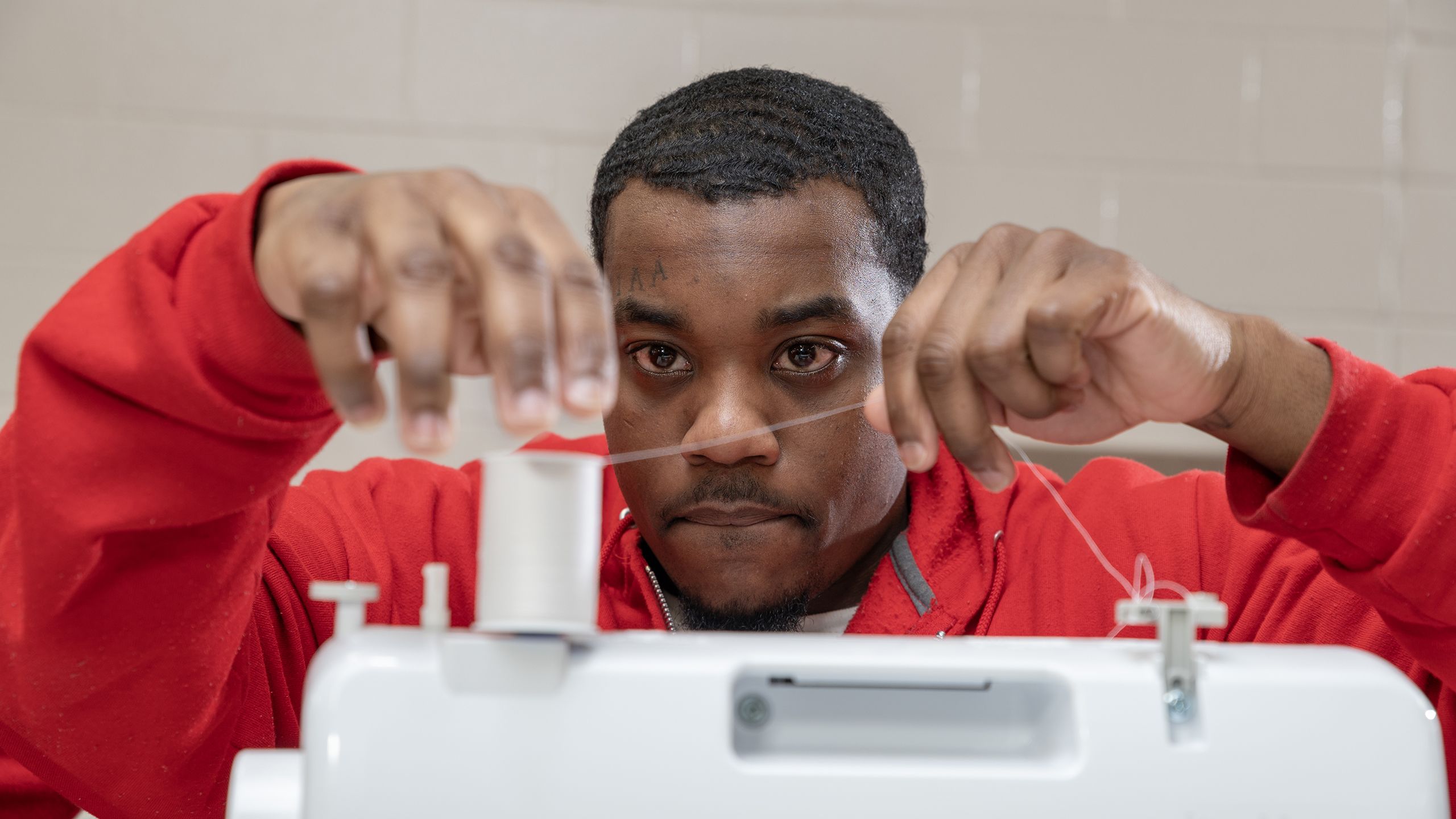 A person intently focusing while threading a needle on a sewing machine. The individual is wearing a red top. The background appears to be a workshop environment, blurred, hinting at a structured setting.