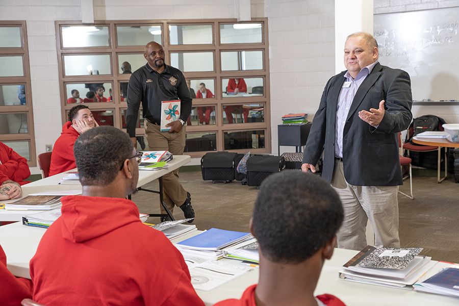 An instructor addressing students in a classroom setting with another person standing aside, surrounded by educational materials and attentive students.