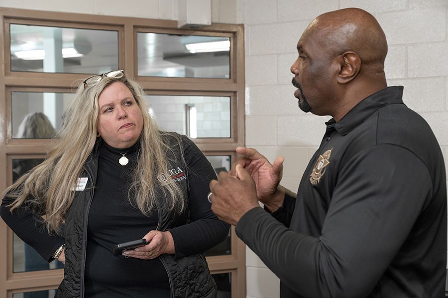 A blonde woman with a UGA jacket listens intently to a sheriff's deputy during a discussion in an office setting.