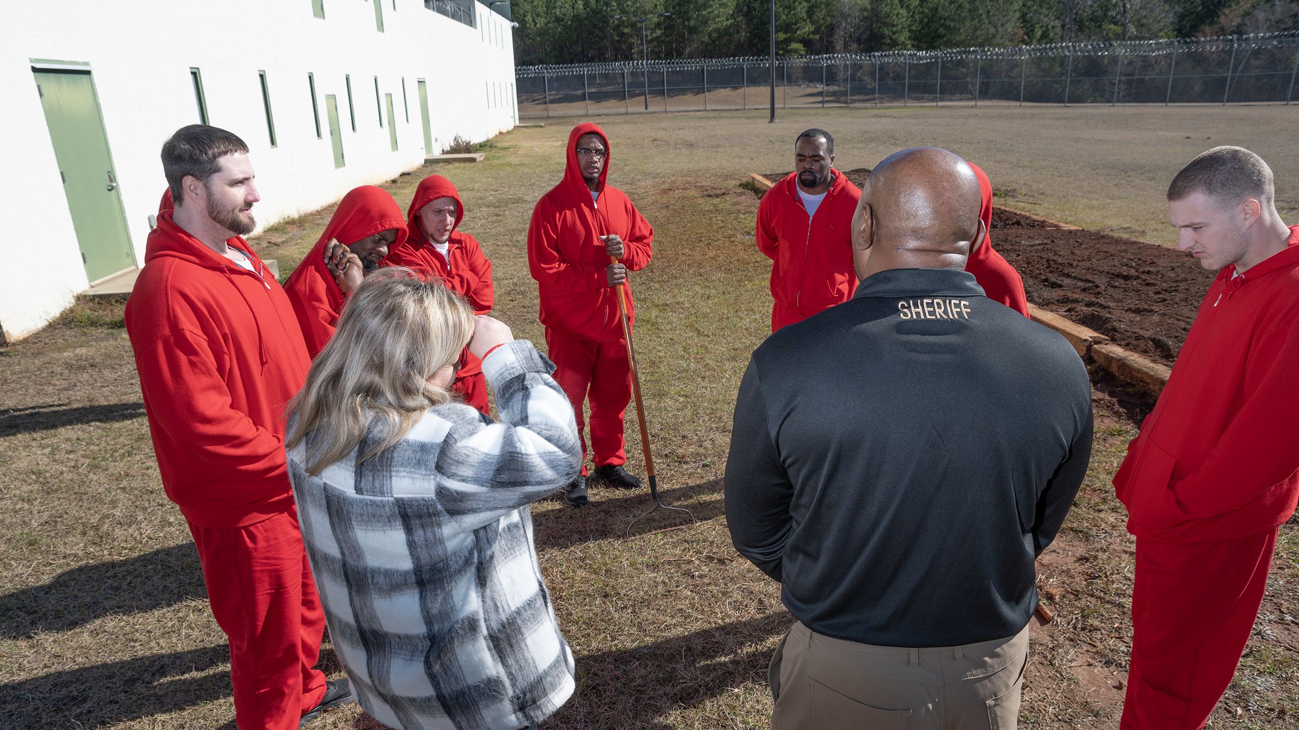 A group of five individuals in red jumpsuits stand outdoors in a grassy area, engaging with two individuals, one wearing a black jacket labeled "SHERIFF" and another in a plaid shirt taking a photo. A building and bare trees are visible in the background, under a clear blue sky.