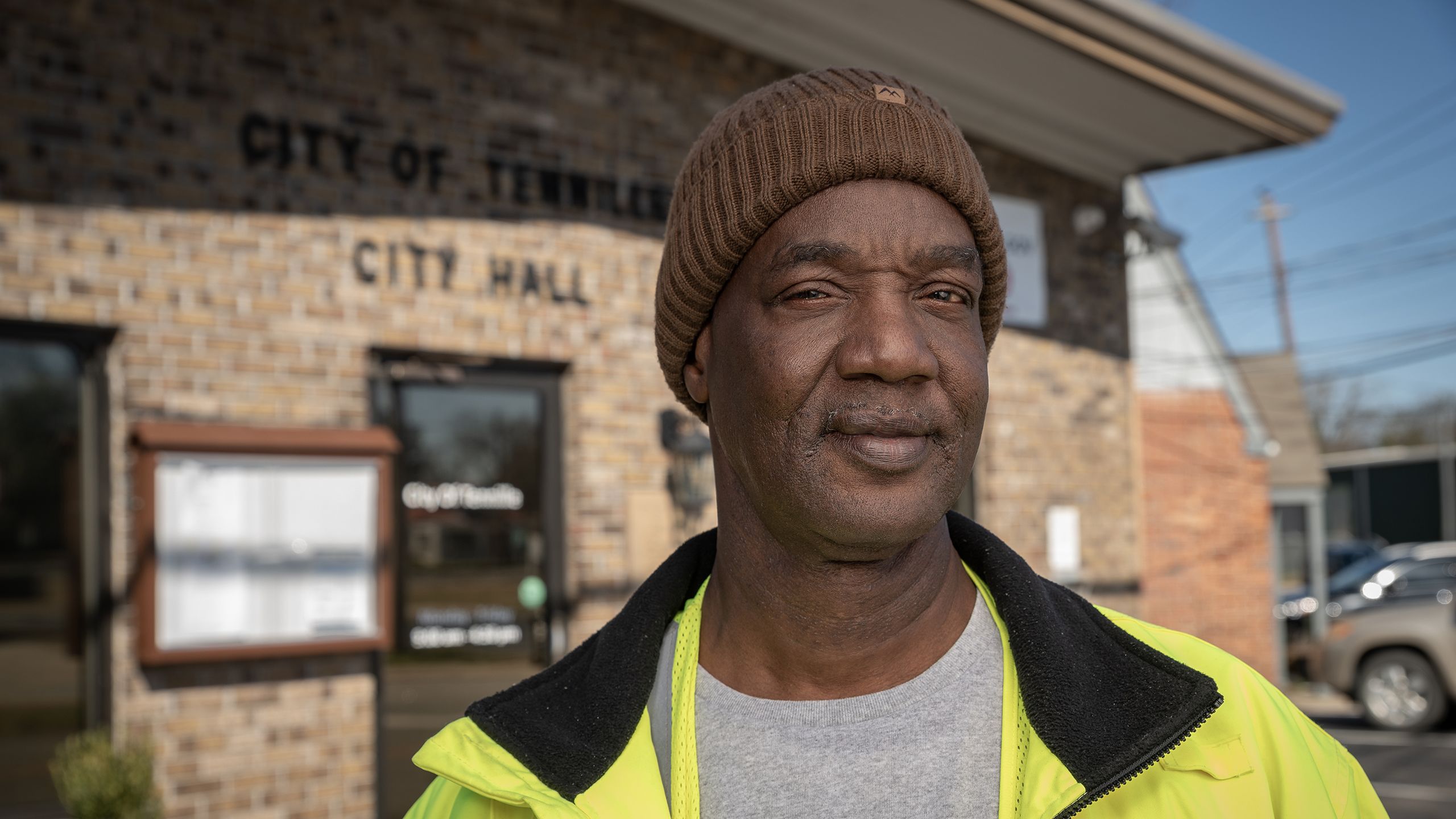 Close-up of a person wearing a neon yellow jacket and brown beanie standing in front of the a city hall building.