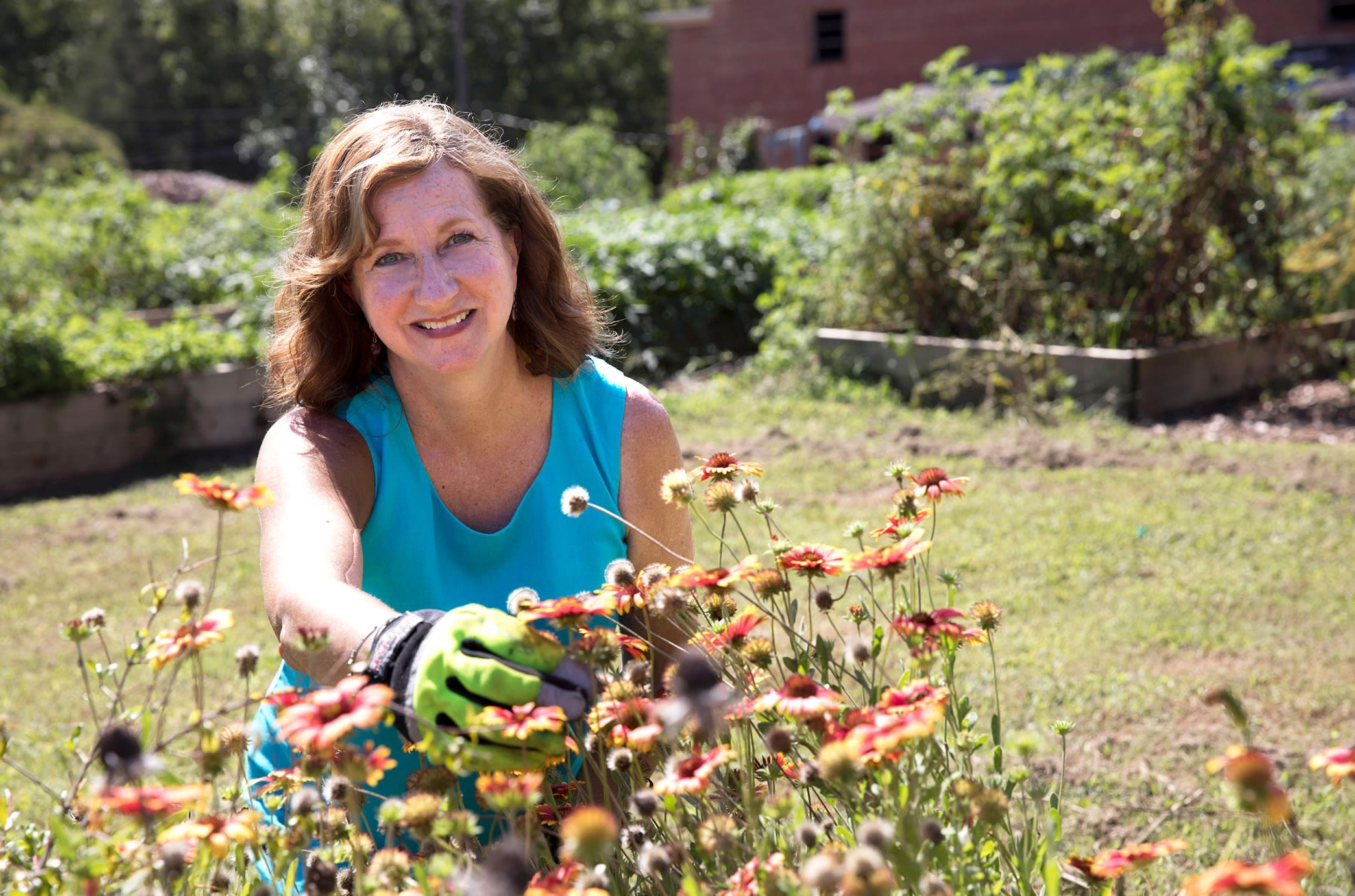 Becky Griffin wears a blue shirt and garden gloves as she touches orange wildflowers in a community garden on a sunny day.
