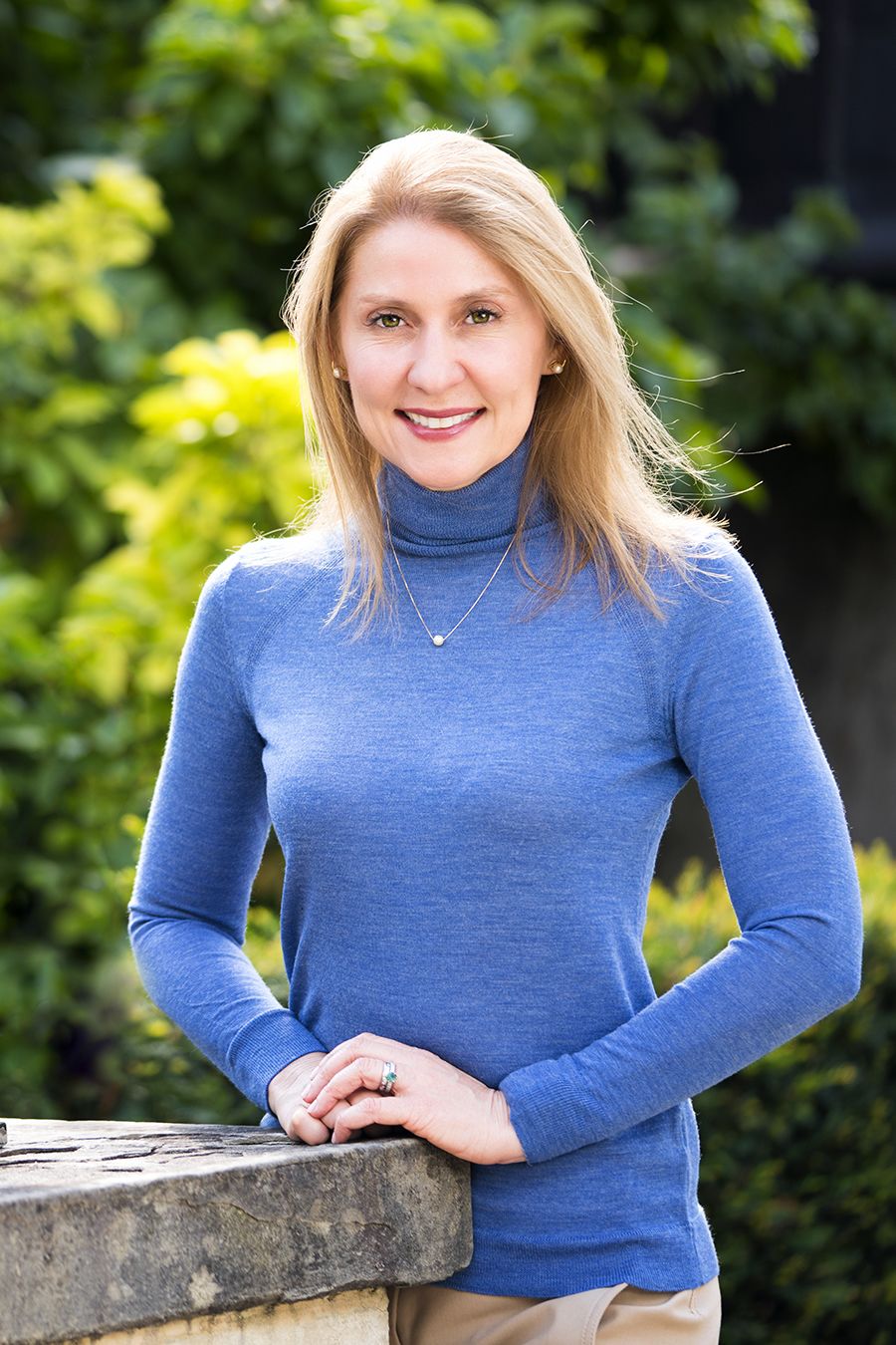 A headshot of a blonde woman wearing a blue sweater against a natural background
