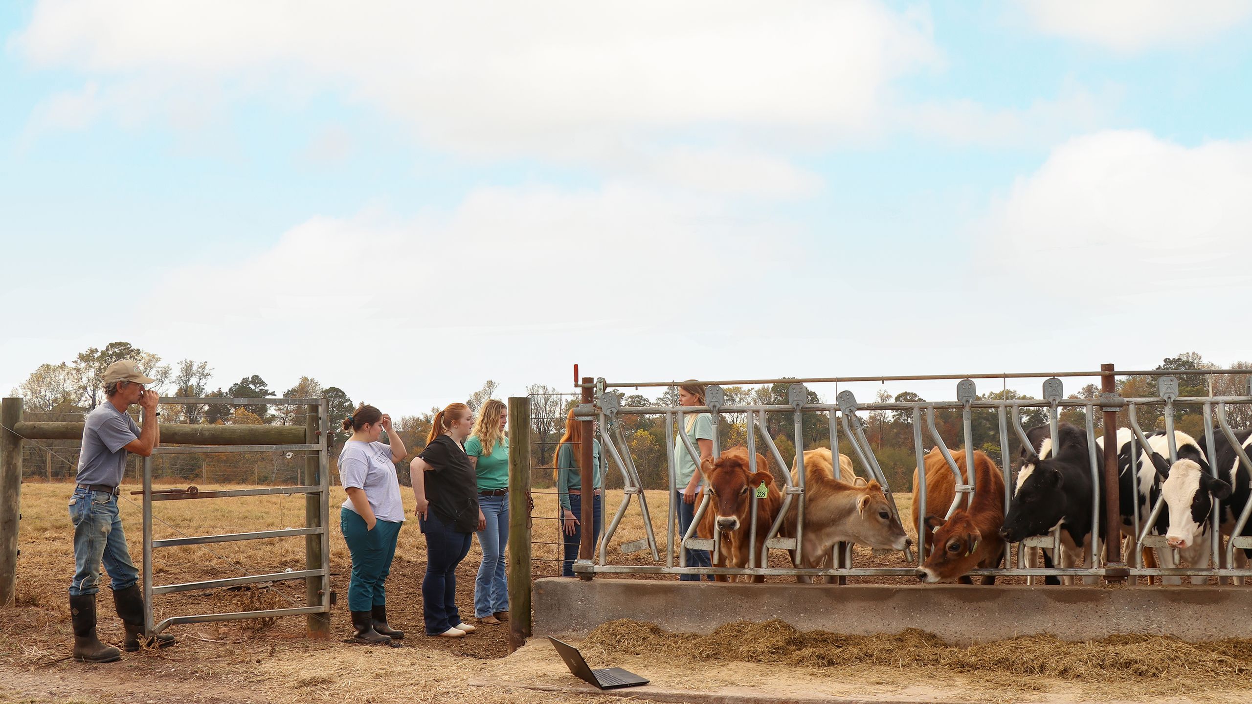 Four people observing cows at a feeding trough, with one person leaning on a gate and another holding agricultural tools, in a rural farm environment.