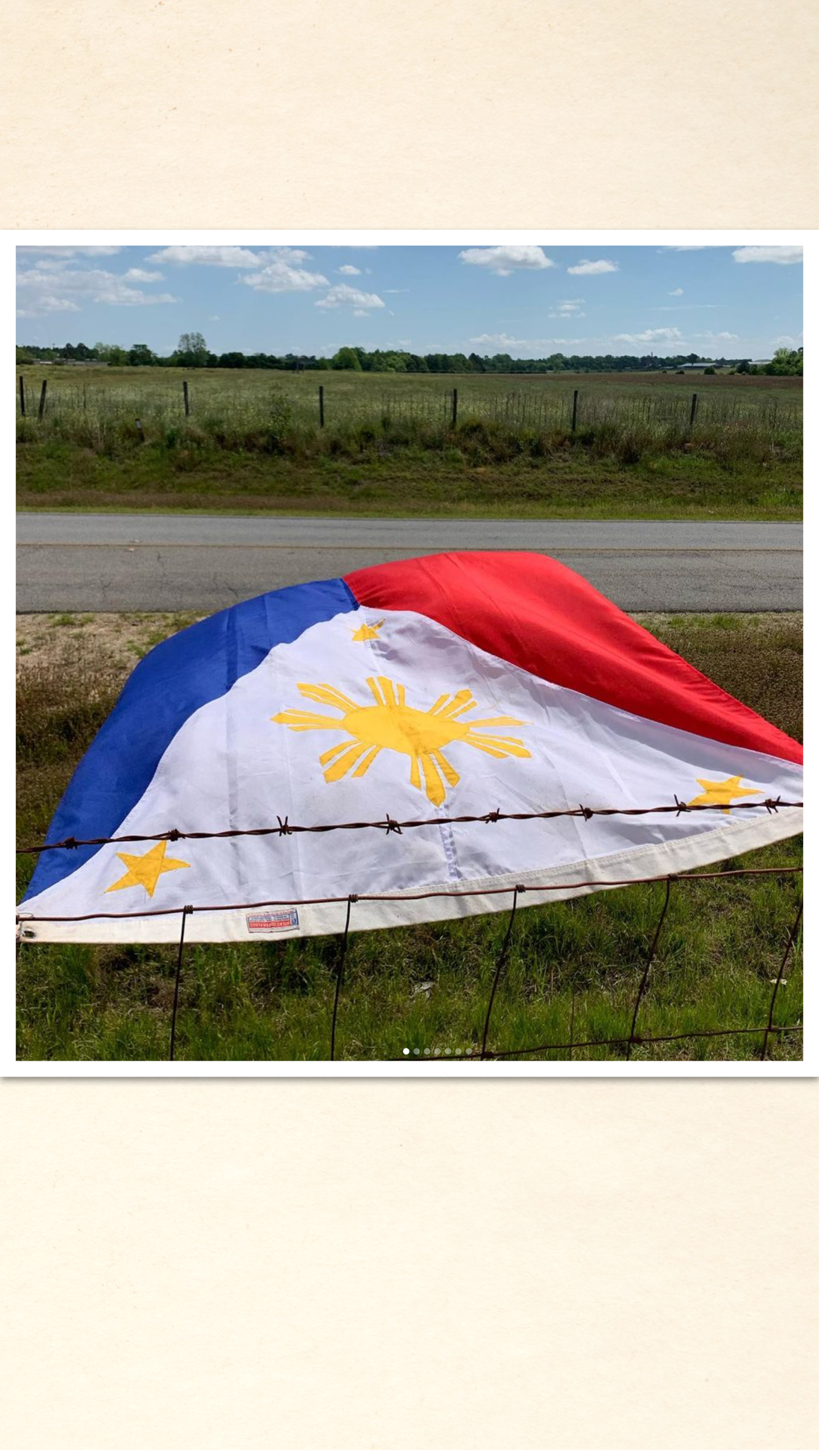 A large, weathered Philippine flag lies on the ground, partially draped over a barbed wire fence with a rural road and open fields in the background.