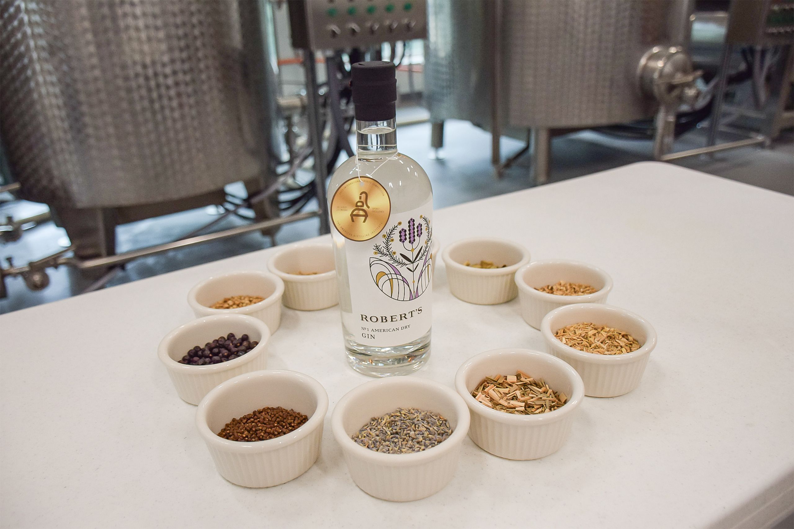 A bottom of gin surrounded by containers filled with ingredients such as lemongrass, cardamom, and more.