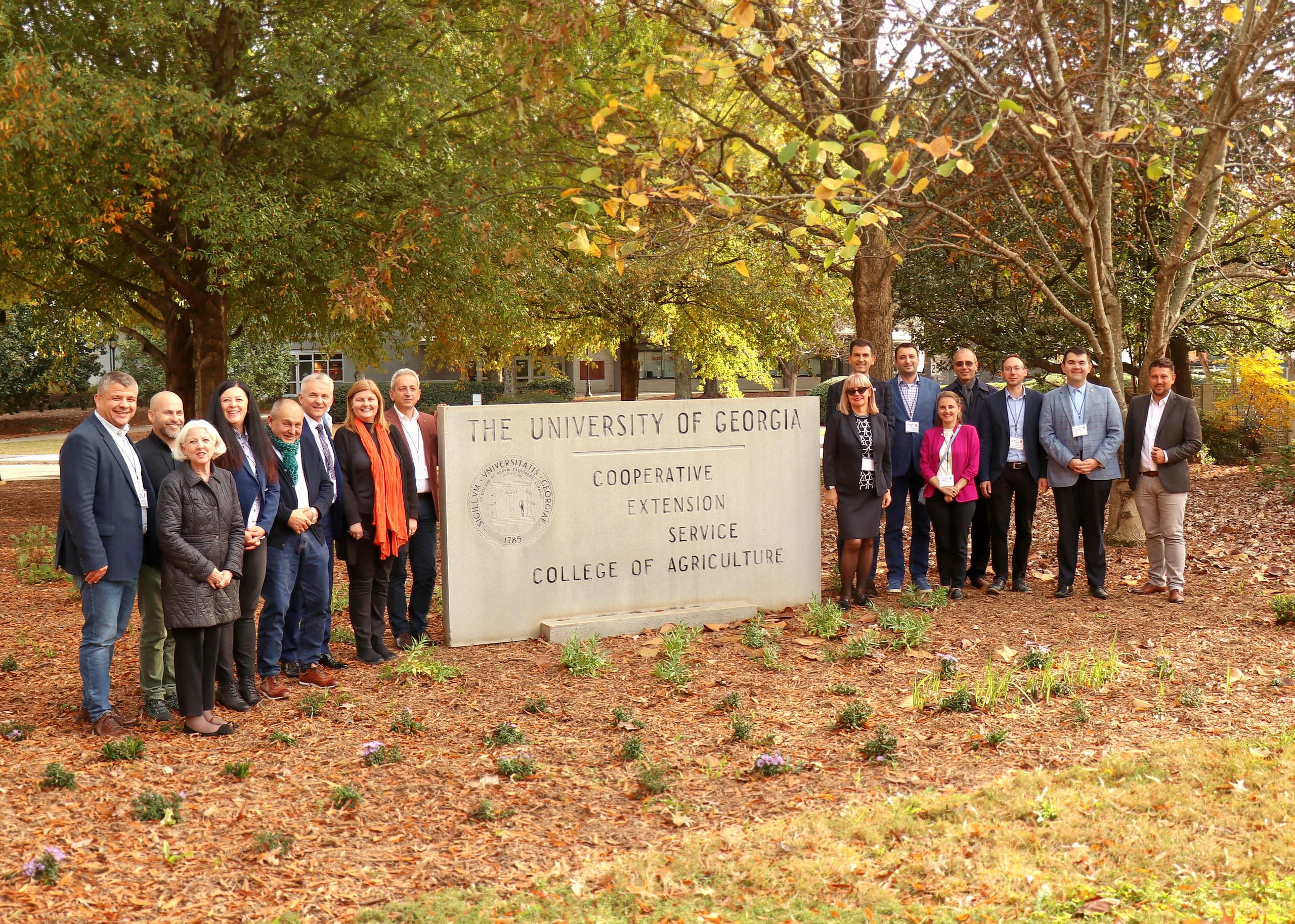 A group of professionally dressed people who are part of the Romanian program stand around a stone sign of The University of Georgia Cooperative Extension Service College of Agriculture on an autumn day with fall foliage in the background.