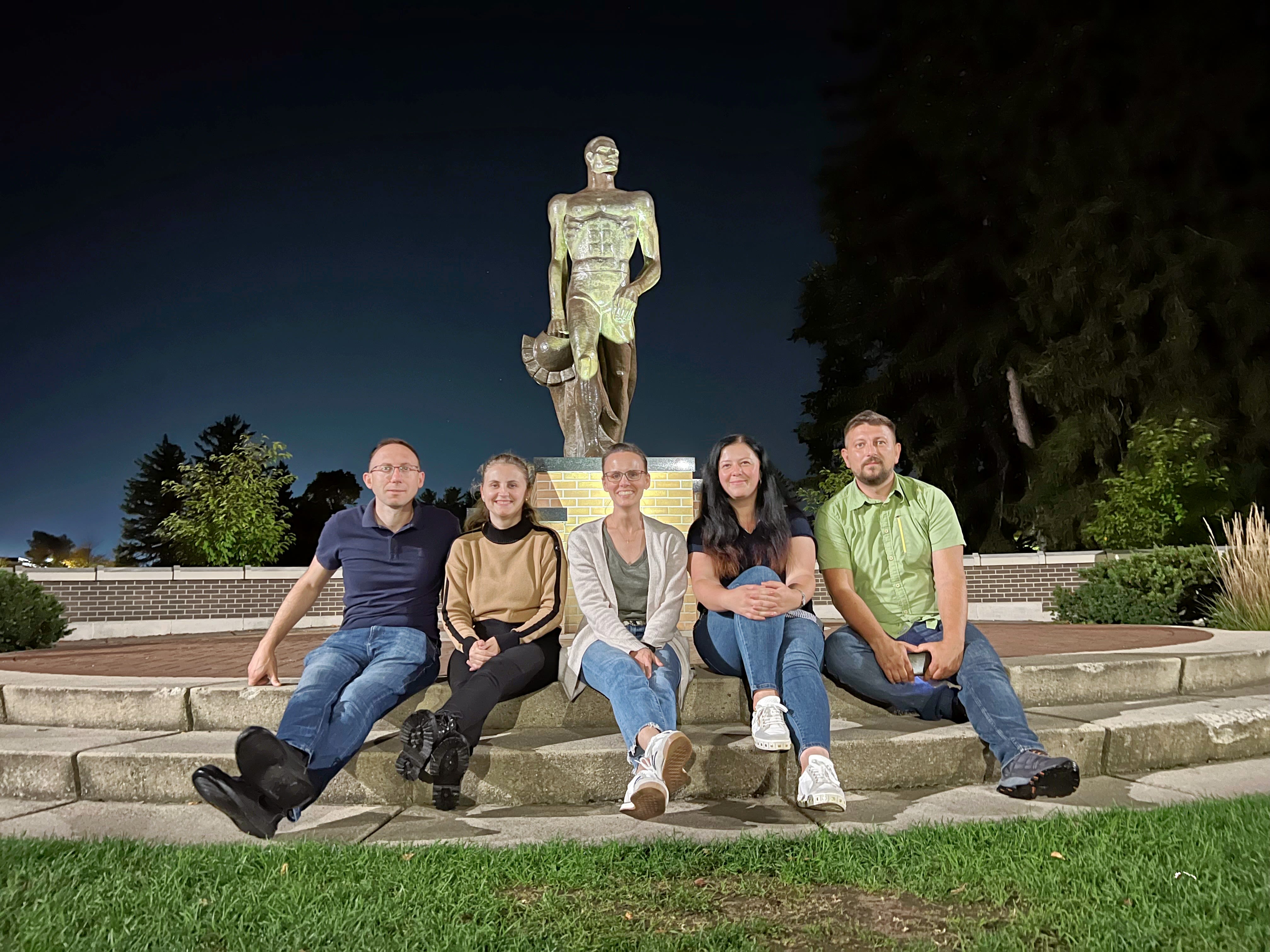 Five people sitting in front of a statue of a roman soldier with night sky in the background. Borron sits in the middle.