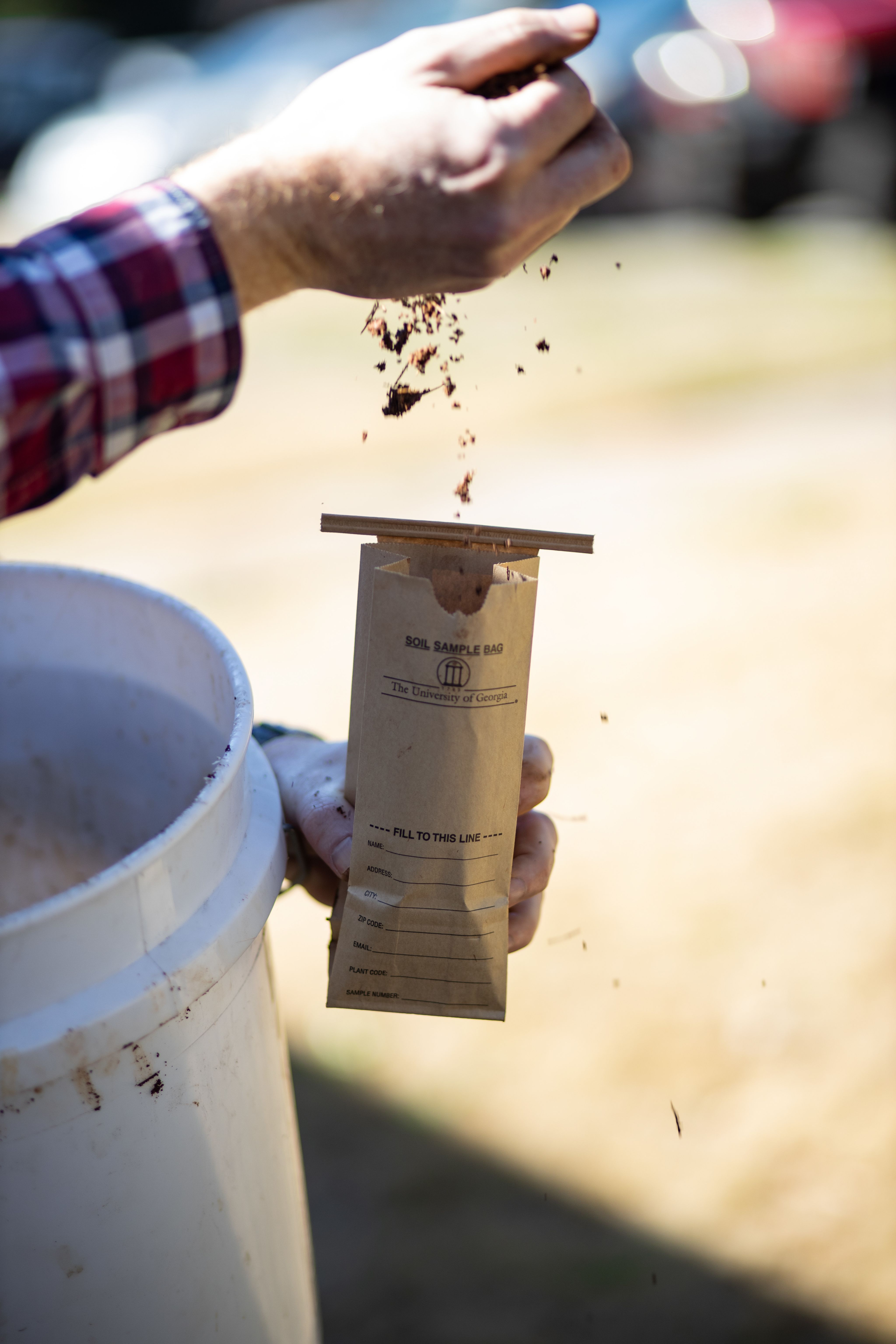 Person holding a brown soil testing bag and white bucket in one arm while filling the bag with soil. 