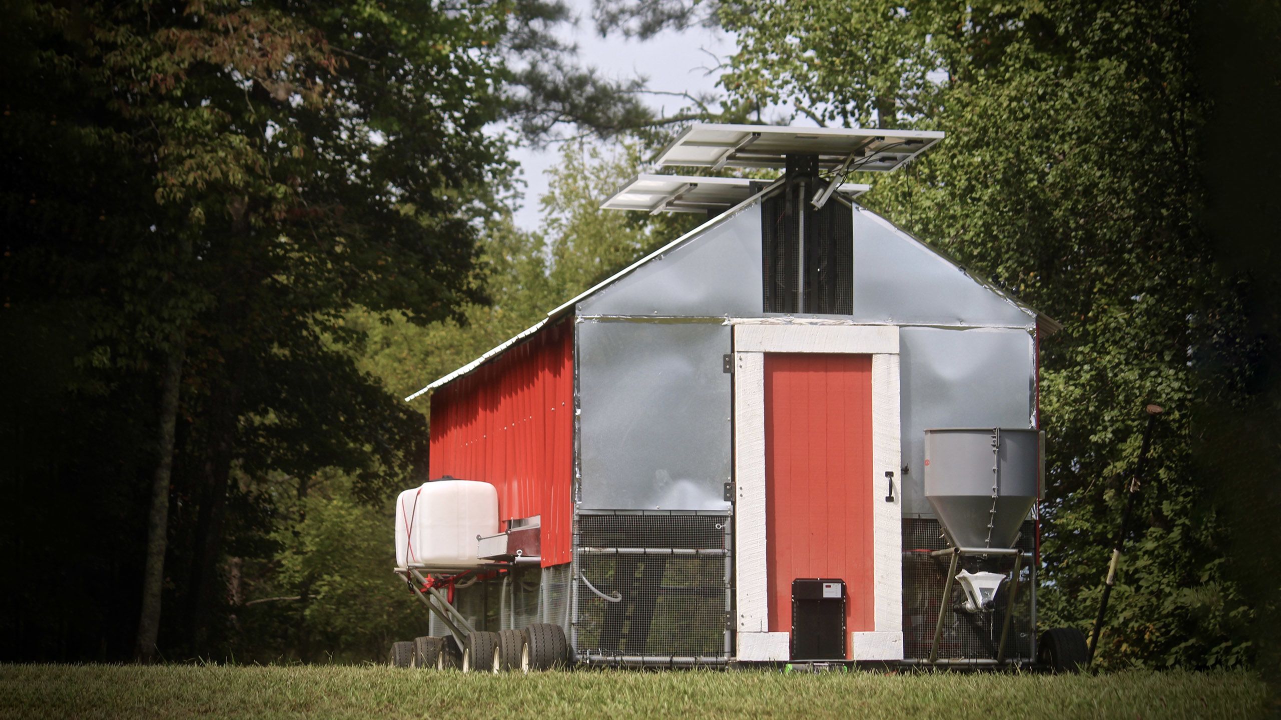 A red and chrome chicken coop with wheels and solar panels sitting in a lush forest scene