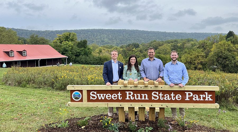 Four people in formal dress standing behind a sign reading "Sweet Run State Park." Behind them is a sprawling mountain landscape.