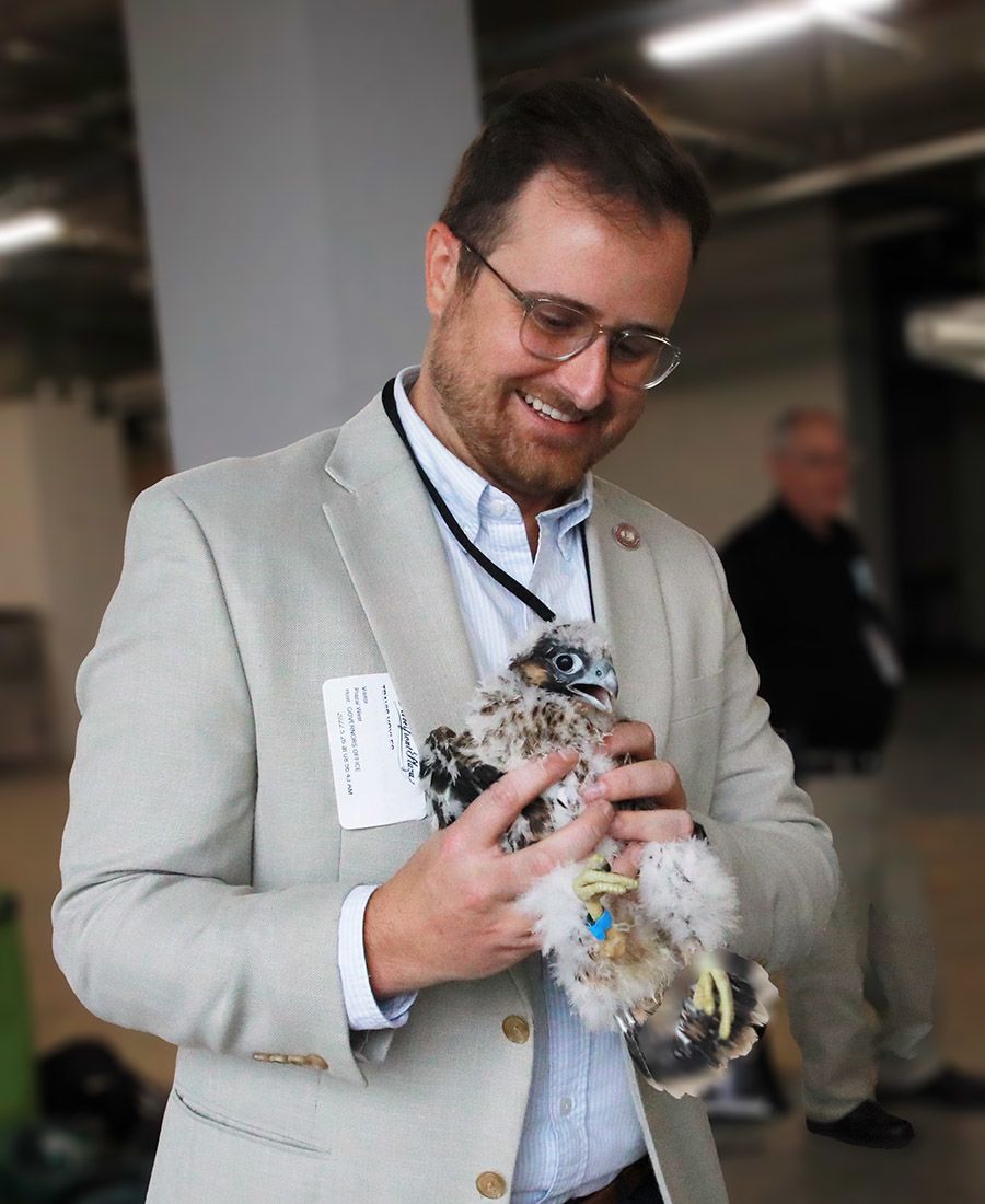 A man in a dress shirt and jacket smiling down at a baby falcon in his hands.