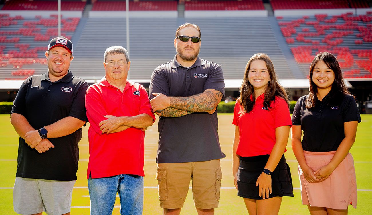 Five people standing side by side in Sanford Stadium. Each wears UGA-affiliated clothing.