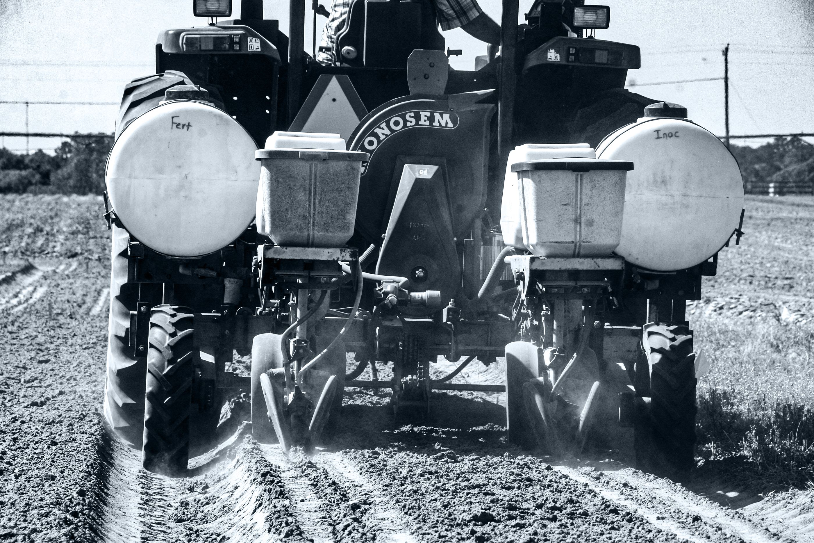Black and white photo of the rear of a tractor carrying canisters of fertilizer and other agricultural chemicals in a newly seeded field.