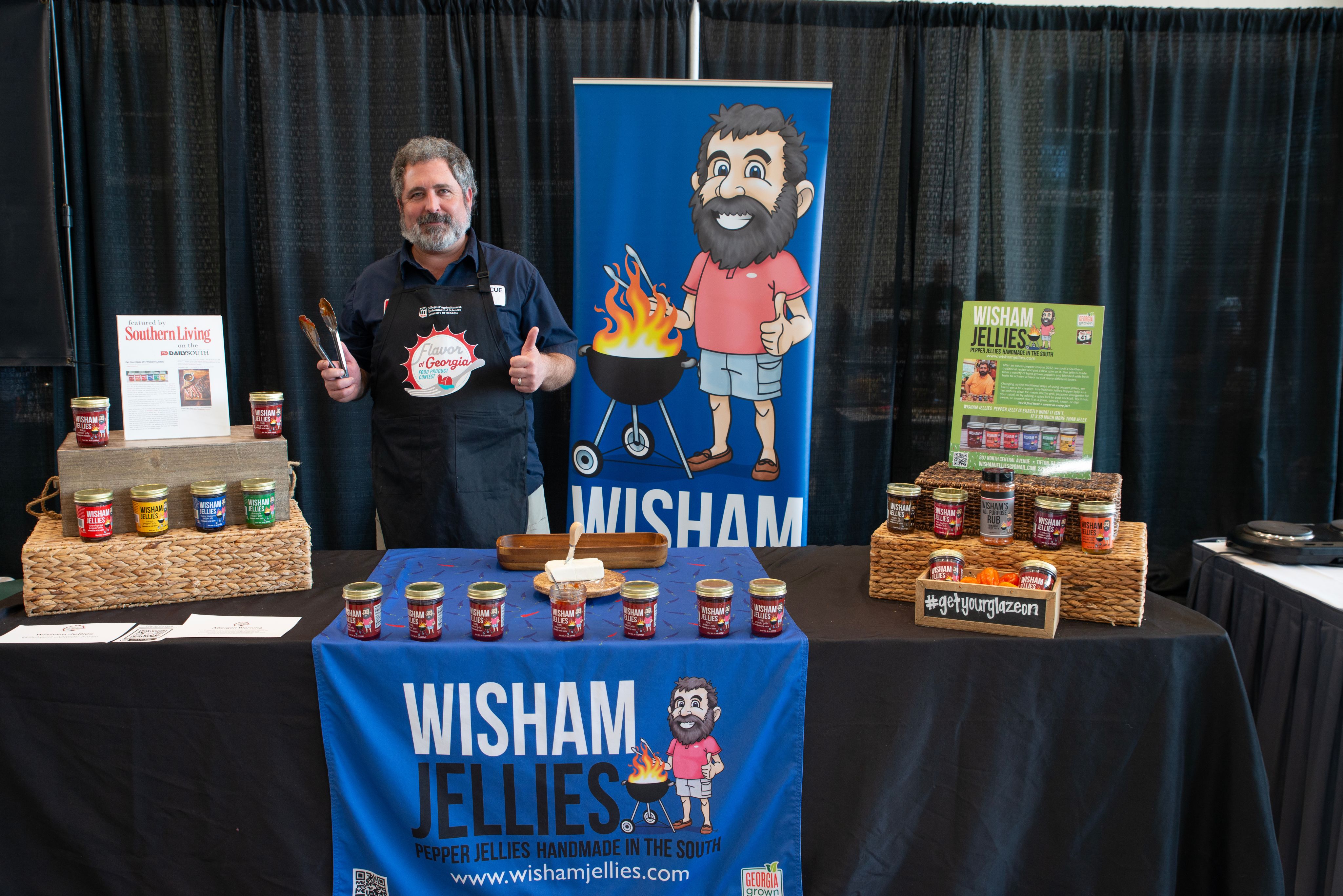 a man in an apron stands behind the Wisham Jellies table, where there are lots of jelly jars displayed in a row and a large blue tablecloth with the wisham jellies logo on it