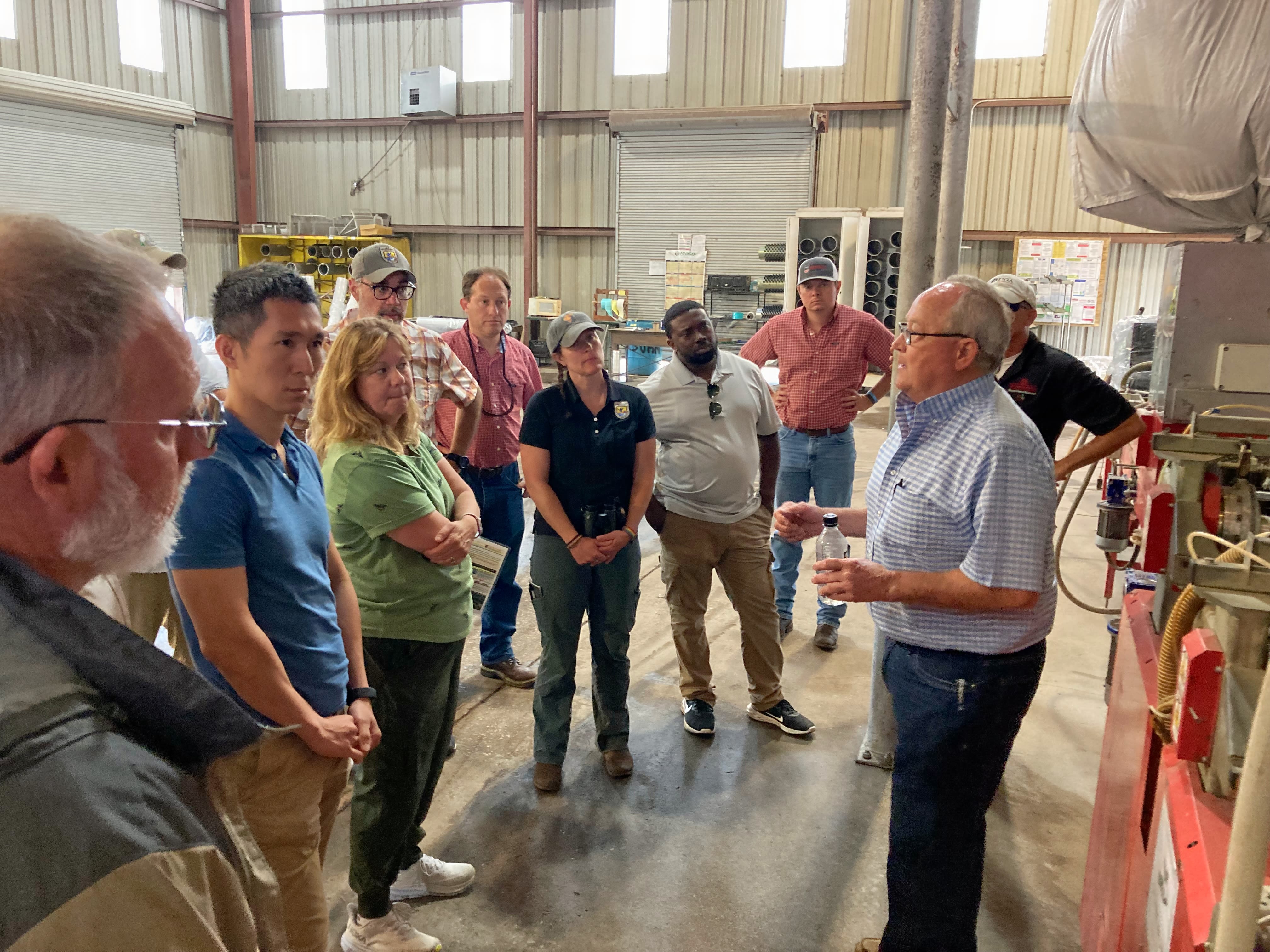 Inside a large packing house, a group of people are gathered around Bill Brim as he talks about the operation. Brim wears a blue and white checkered shirt and glasses.