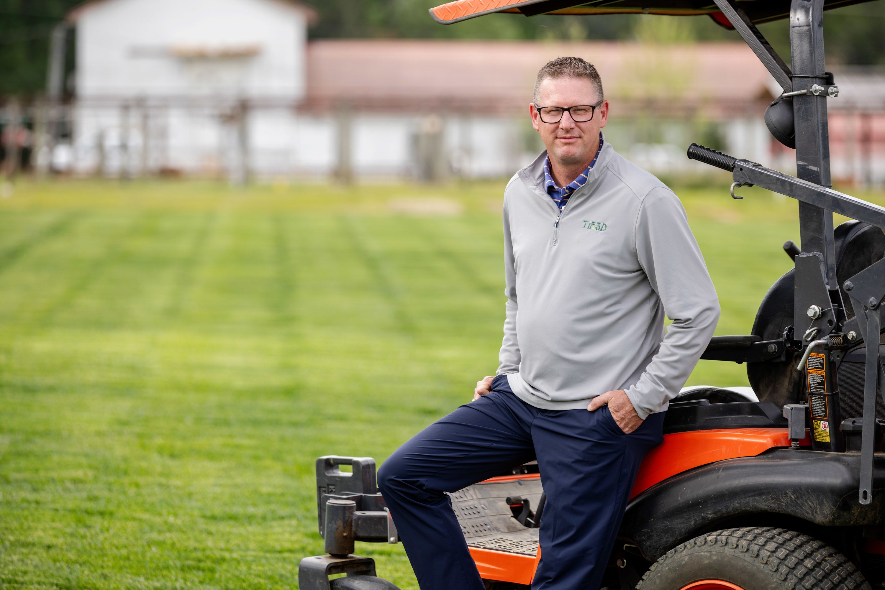 Brian Schwartz sits on the back of a lawn mower at the turfgrass research plots. Trials of green turfgrass can be seen in the background.