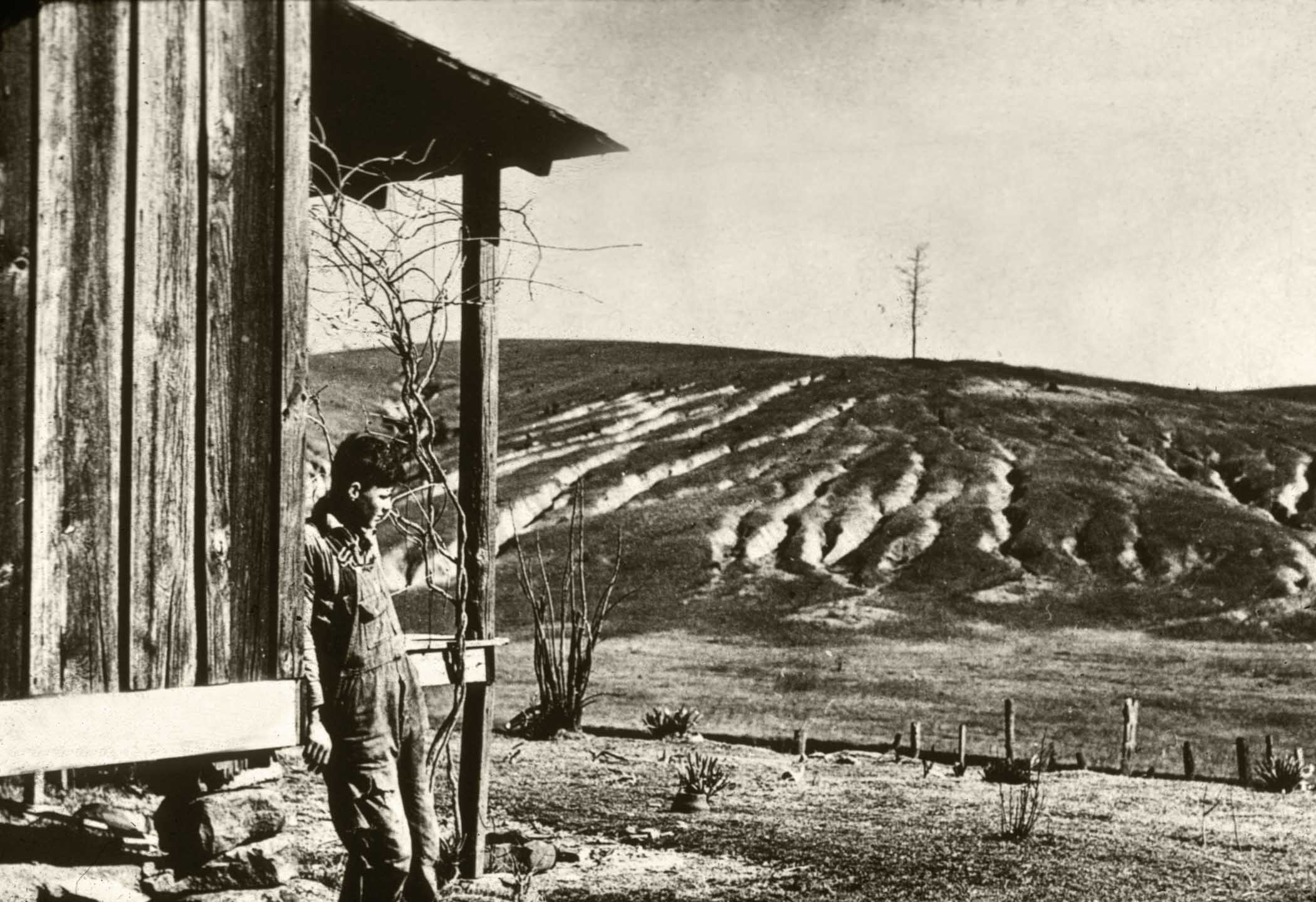 A historical black and white photo of a farmer standing on the porch of a weather farm building with acres of eroded farmland and skeletal trees in the background.