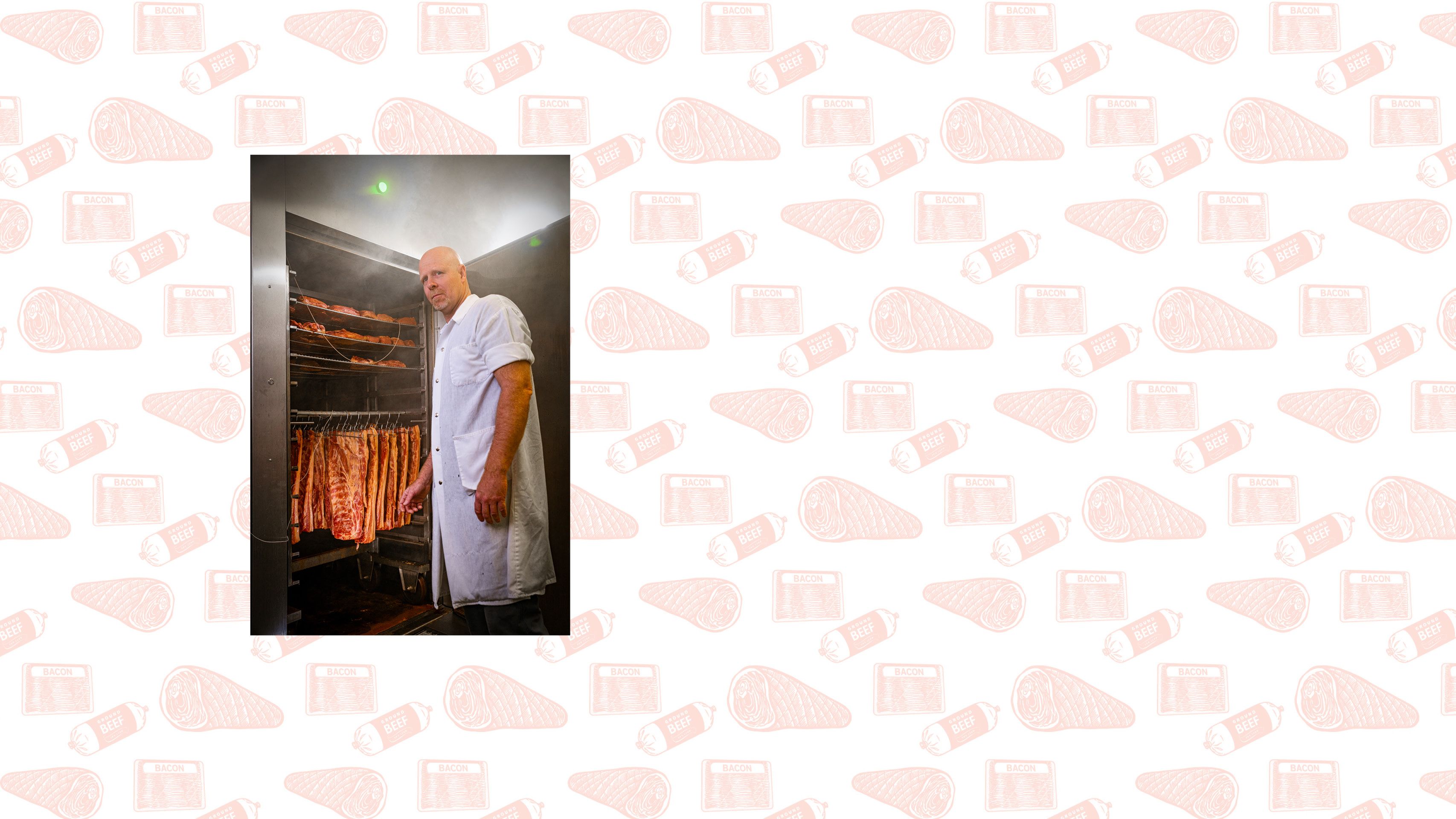 A white man in a white coat stands in front of the open door of a meat smoker. Inside are several racks of meat being smoked.