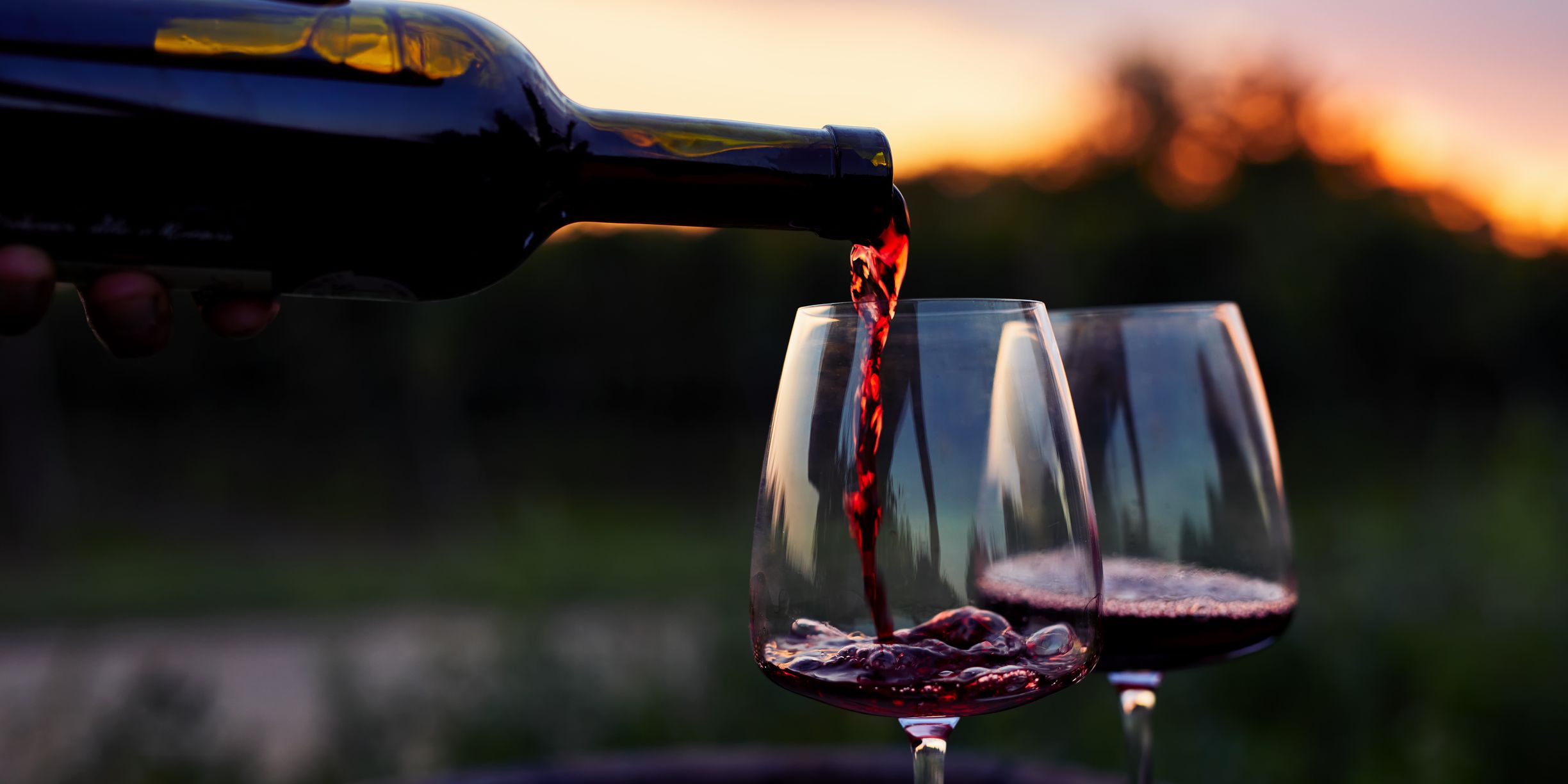 Sunlight glints through red wine poured from a bottle into two glasses