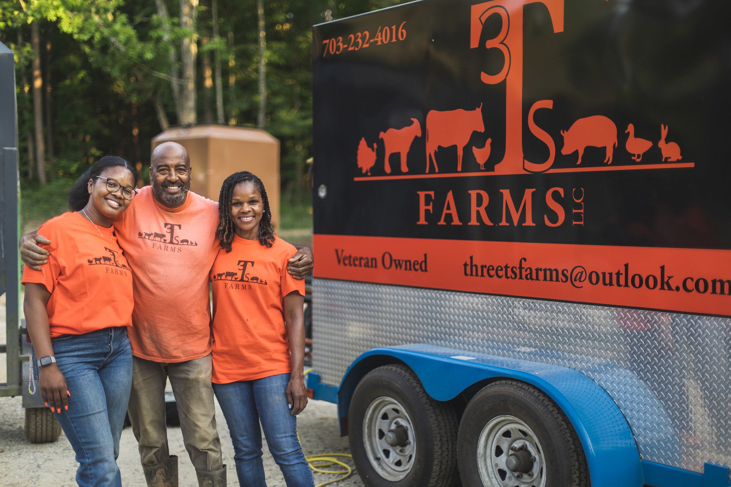 Iteago Felton, his wife, Antheena, and their daughter, Anteaona are all wearing orange t-shirts and standing next to a trailer that says T Farms, Veteran Owned on the side. 