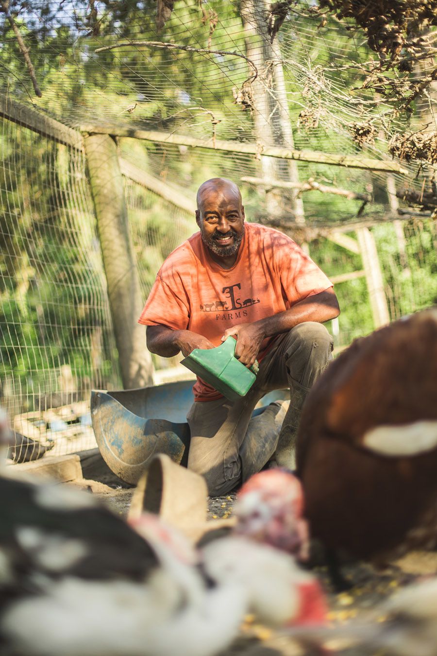 Iteago Felton, kneeling down to feed some animals. In his hand, he's holding a green scoop full of feed. In the foreground are the animals he is feeding, including a duck and a turkey.