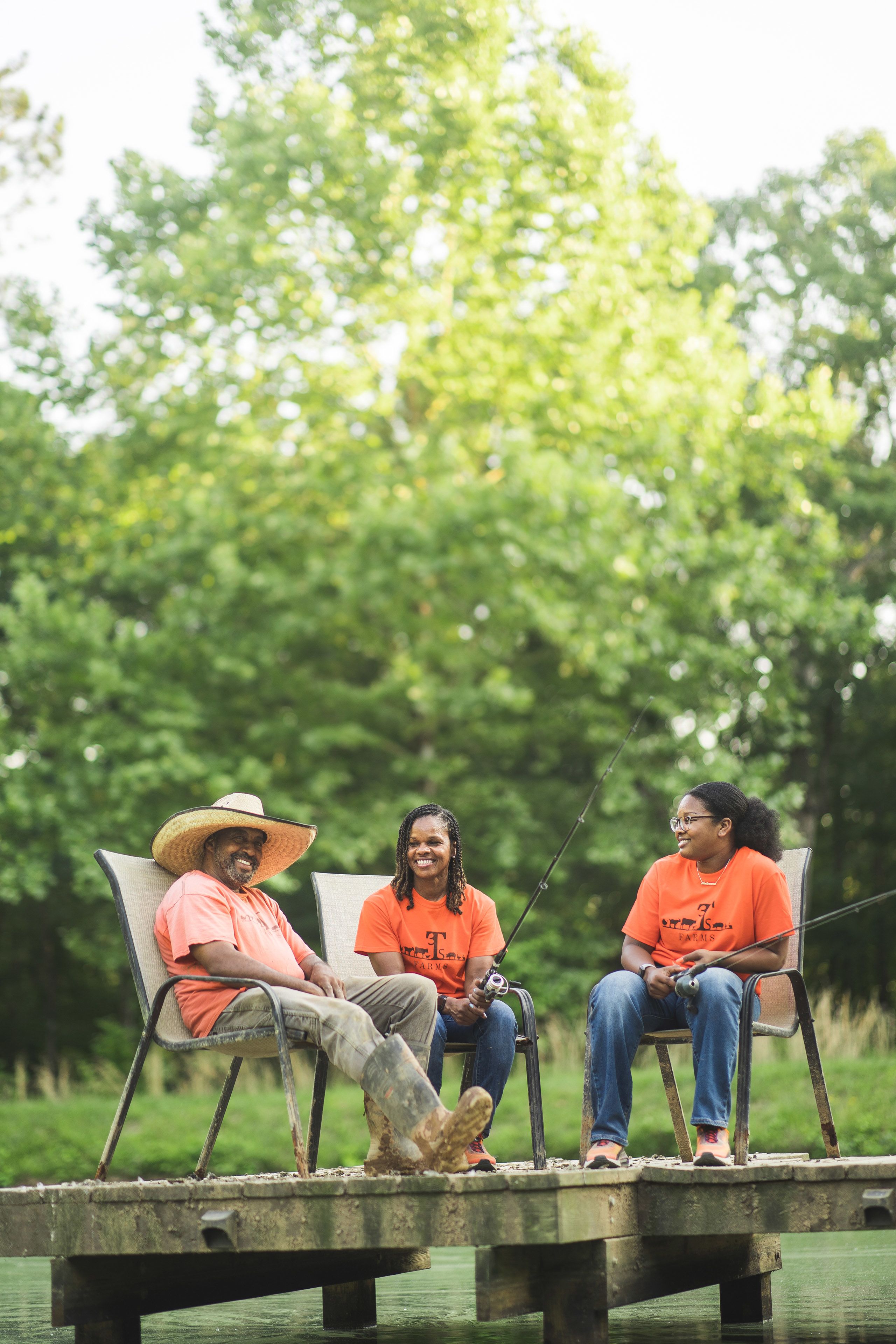 Iteago Felton, his wife, Antheena, and their daughter, Anteaona sitting in chairs. They are all wearing orange t-shirts. Iteago is wearing a large straw hat. The women are both holding fishing poles.