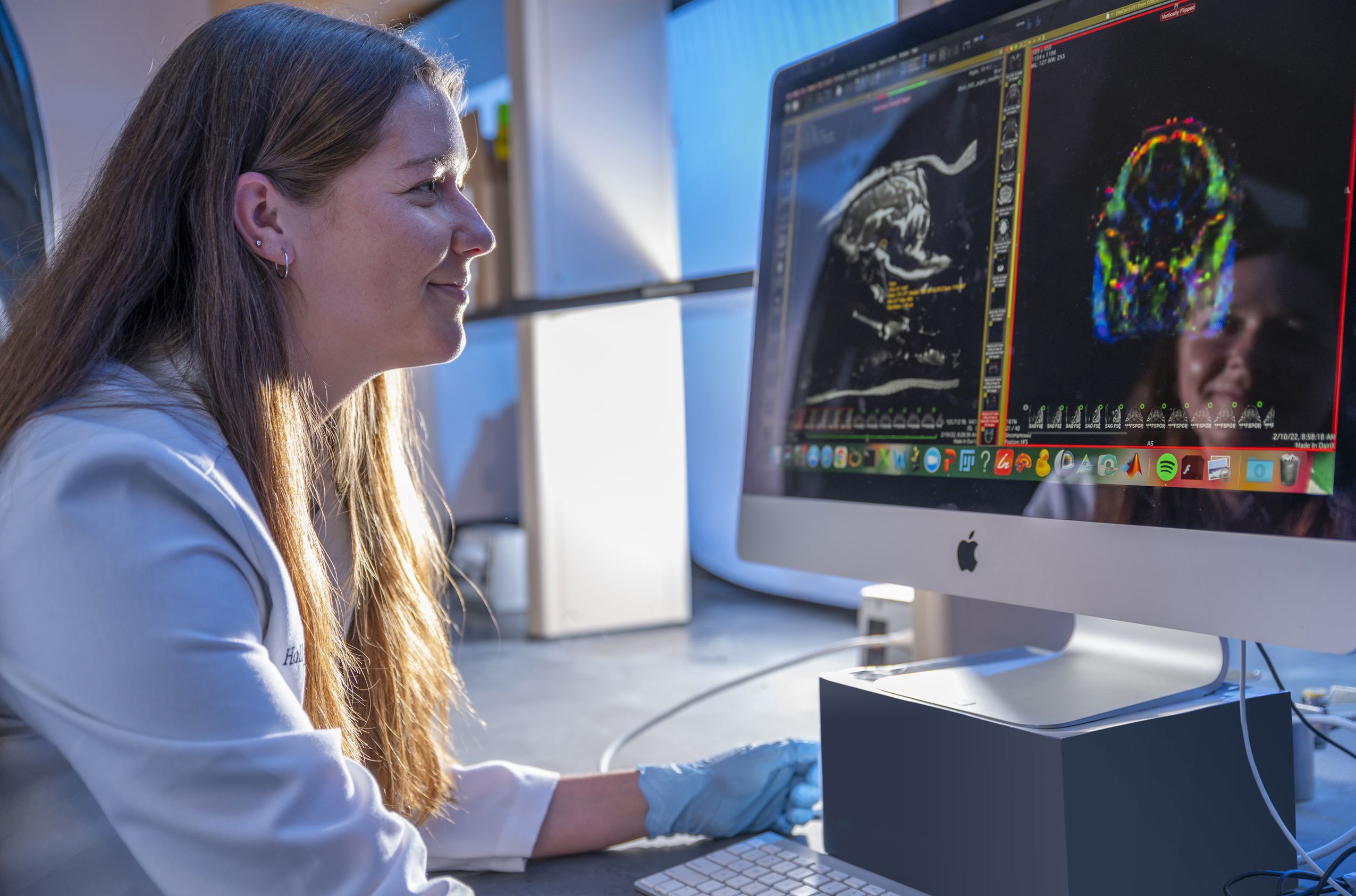 A student wearing a lab coat and gloves looks at side-by-side scans of a pig brain on a computer monitor.