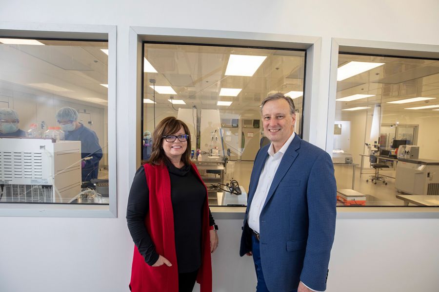 Steven Stice and his wife, Tracey Stice, stand in front of a bank of windows looking into an RBC lab. Steven Stice s wearing a blue suit and Tracey Stice wears a black outfit with a long red vest and glasses. 