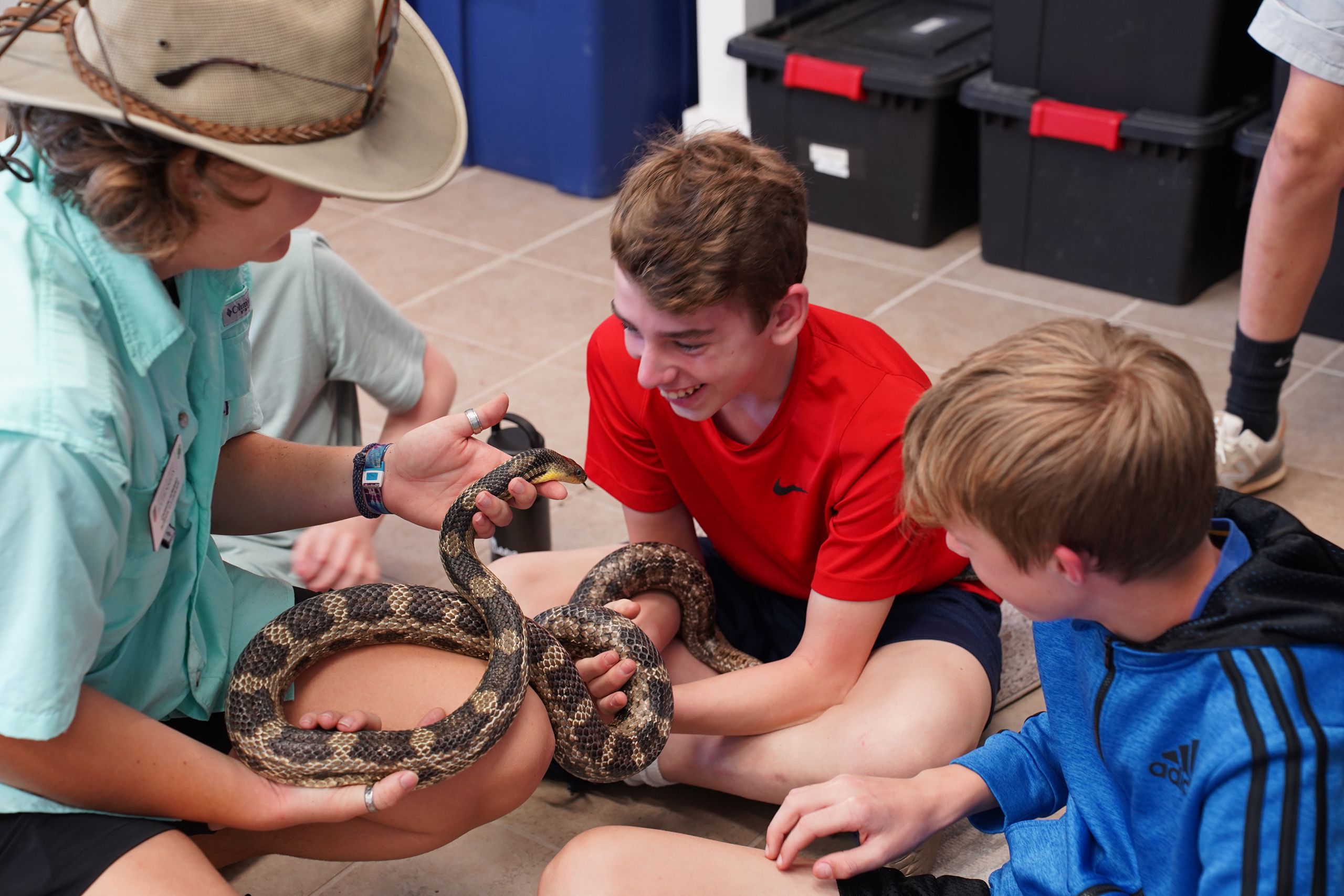 A 4-H educator sits on the ground with two 4-H students, holding a large snake.