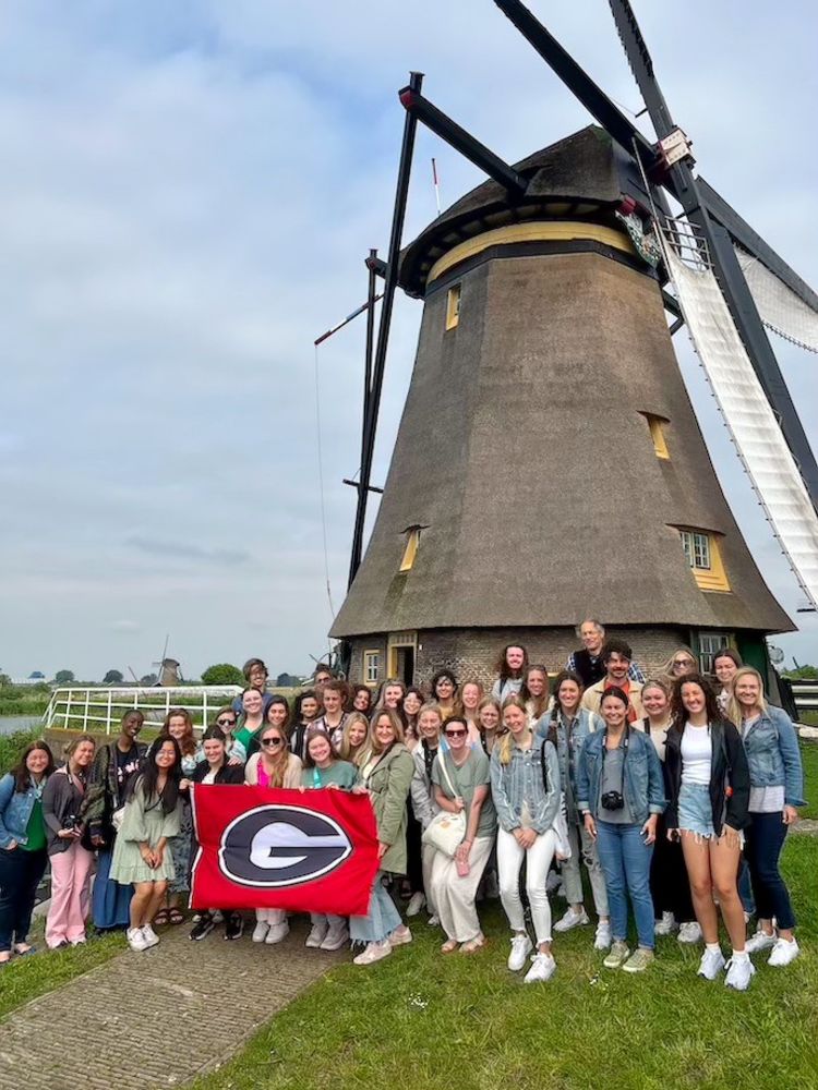 The students hold a UGA flag in a large group photo in front of a Dutch windmill