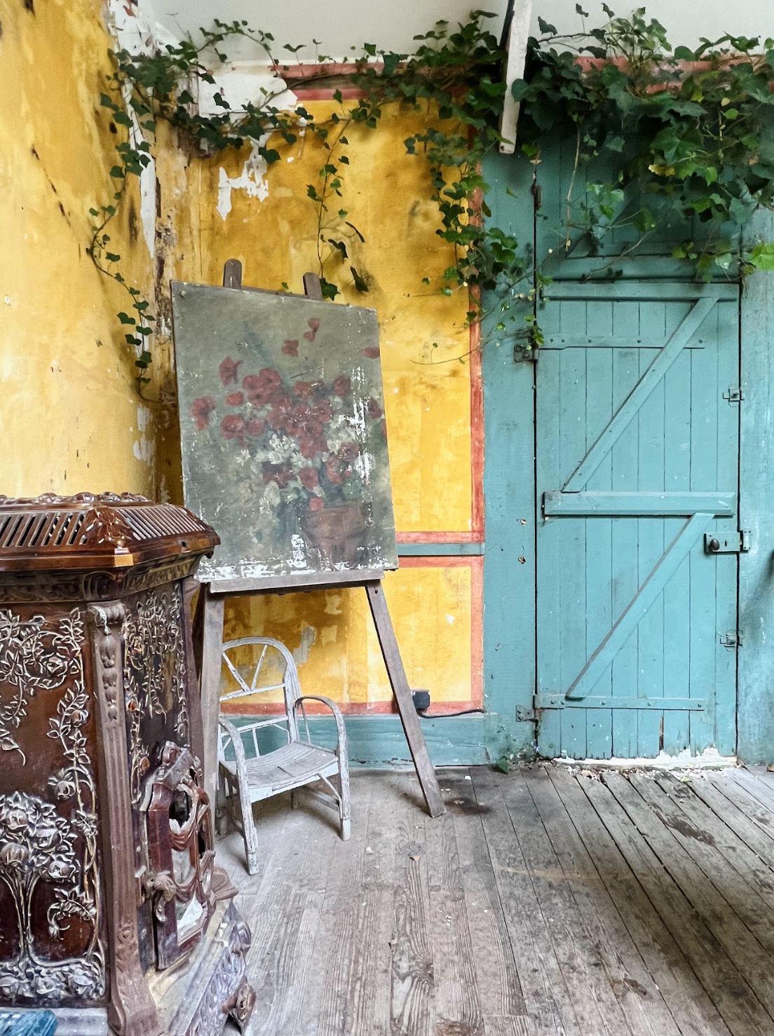 A painting of a bouquet is displayed outside a turquoise door with vines growing up the yellow exterior walls