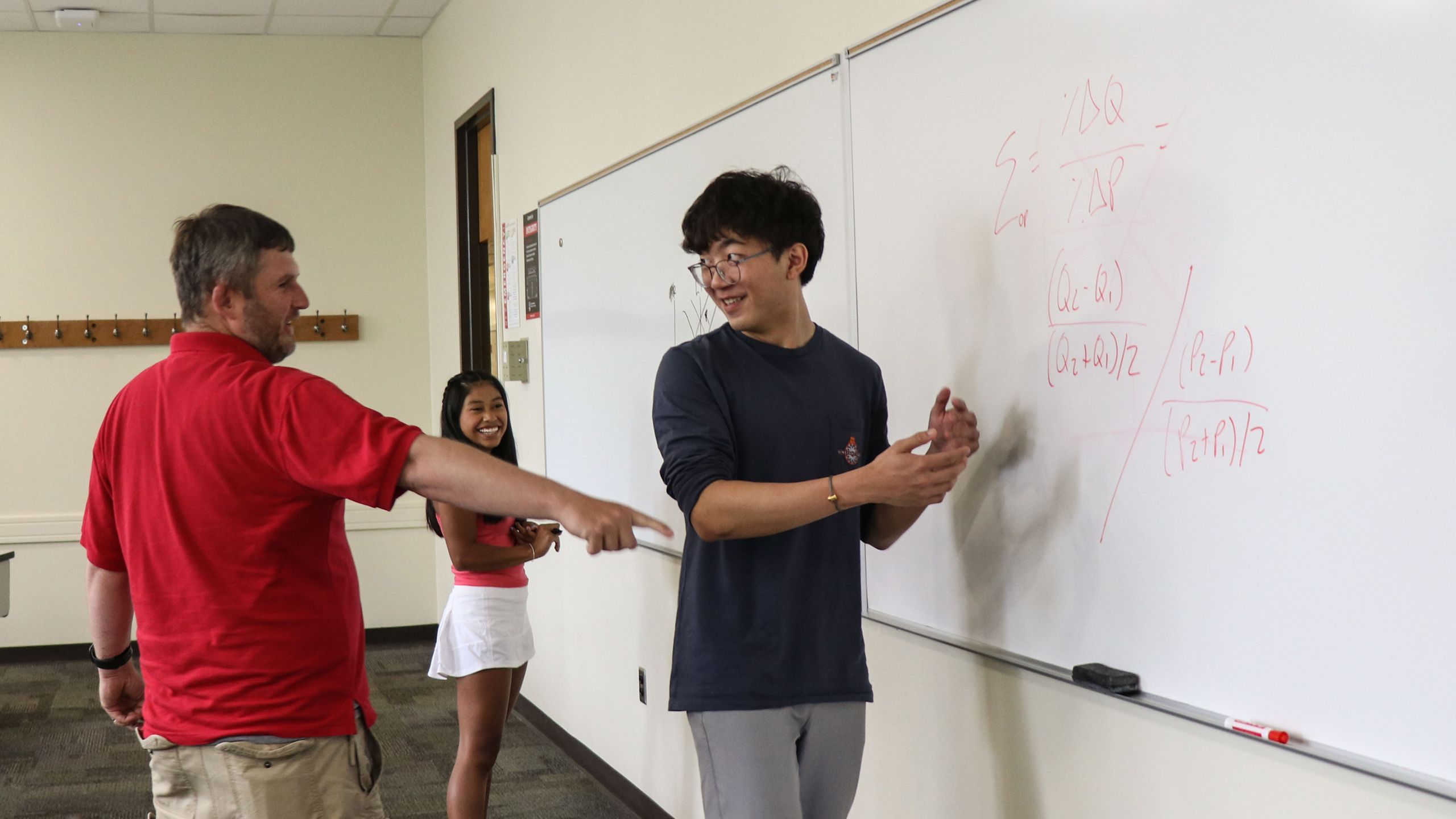 Maya Brantley, Nicholas Xu, and Ben Campbell stand at a whiteboard with economic equations written on it.