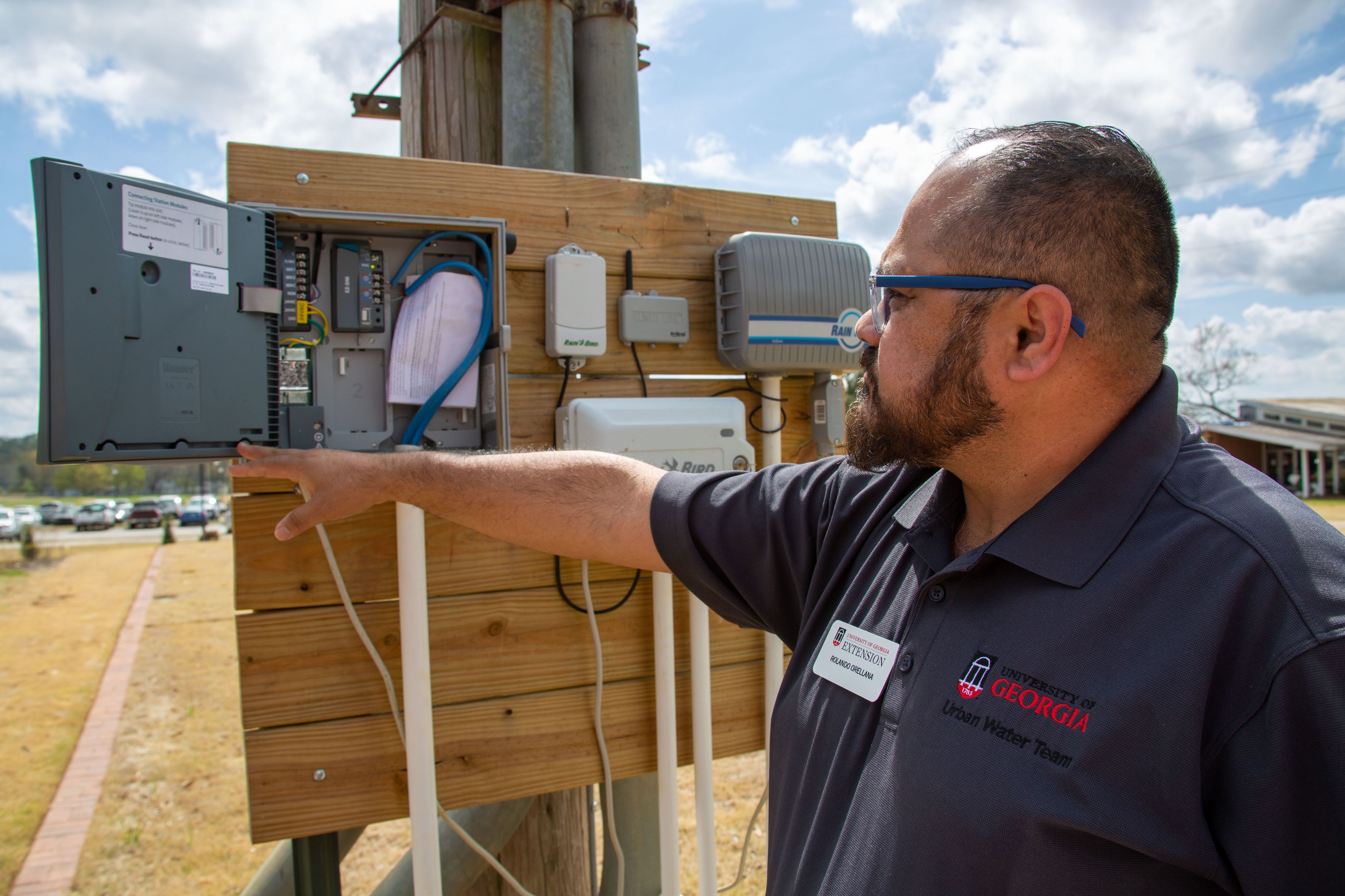 A close up picture of Orellana, whose wearing glasses and a black shirt. He's pointing to a control panel with various water sprinkler control systems.