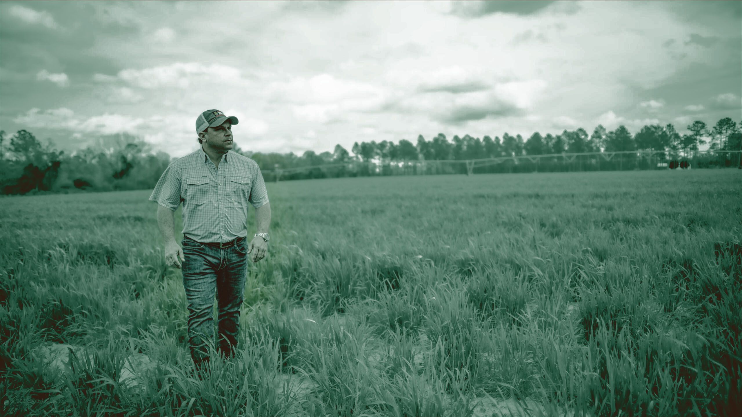 Tyler Harper walks through a grassy field wearing work clothes. The image is overlaid with a dull green filter.