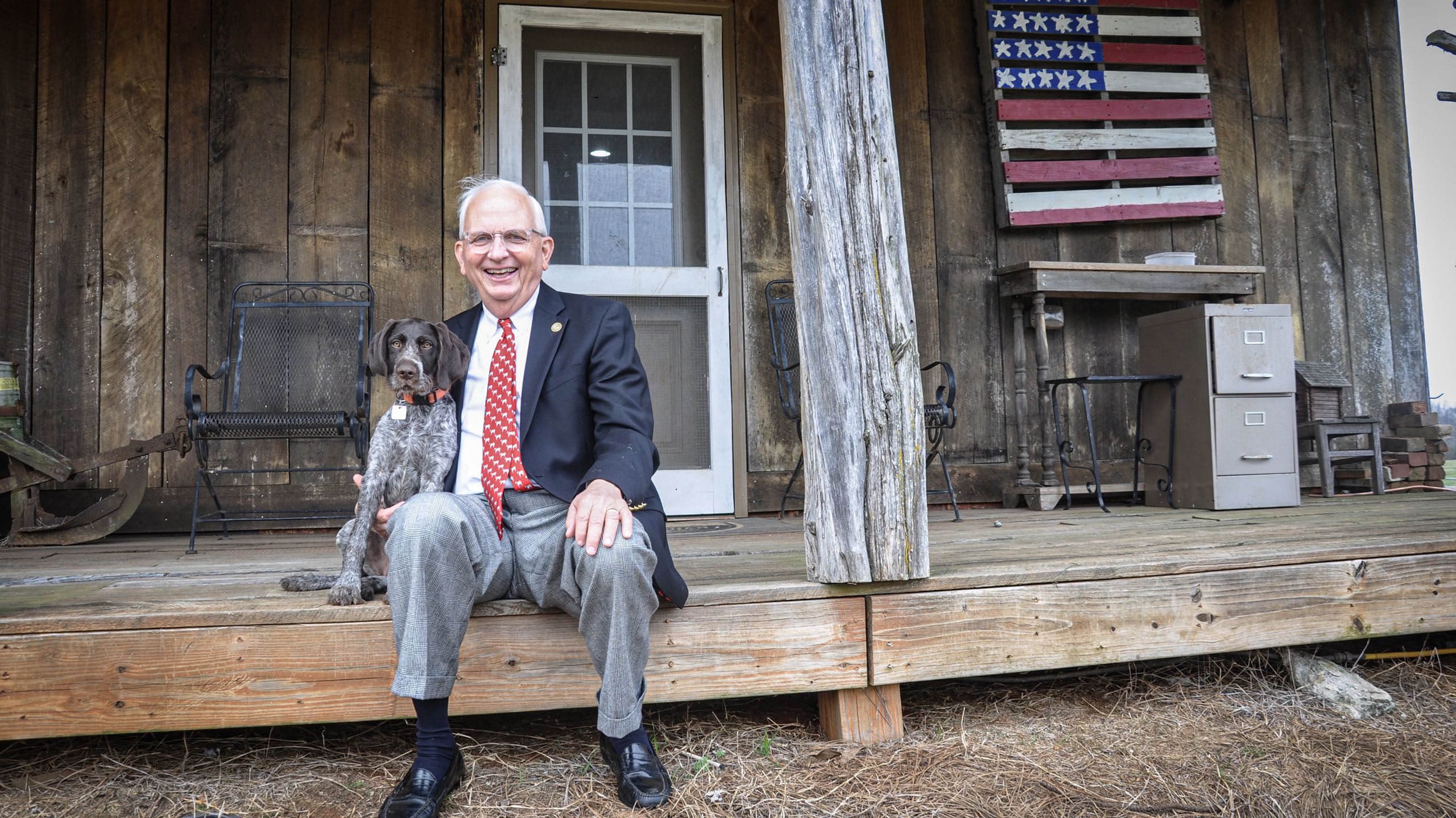 Gary Black sits on a porch with his arm around a pointer dog sitting next to him.