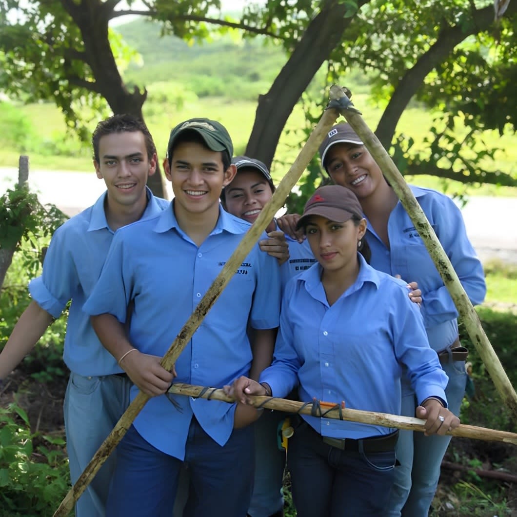 Five UNAG students wearing their blue shirt and blue jean uniforms hold a bamboo A-frame which frames Alejandra. Trees and green fields are in the background.