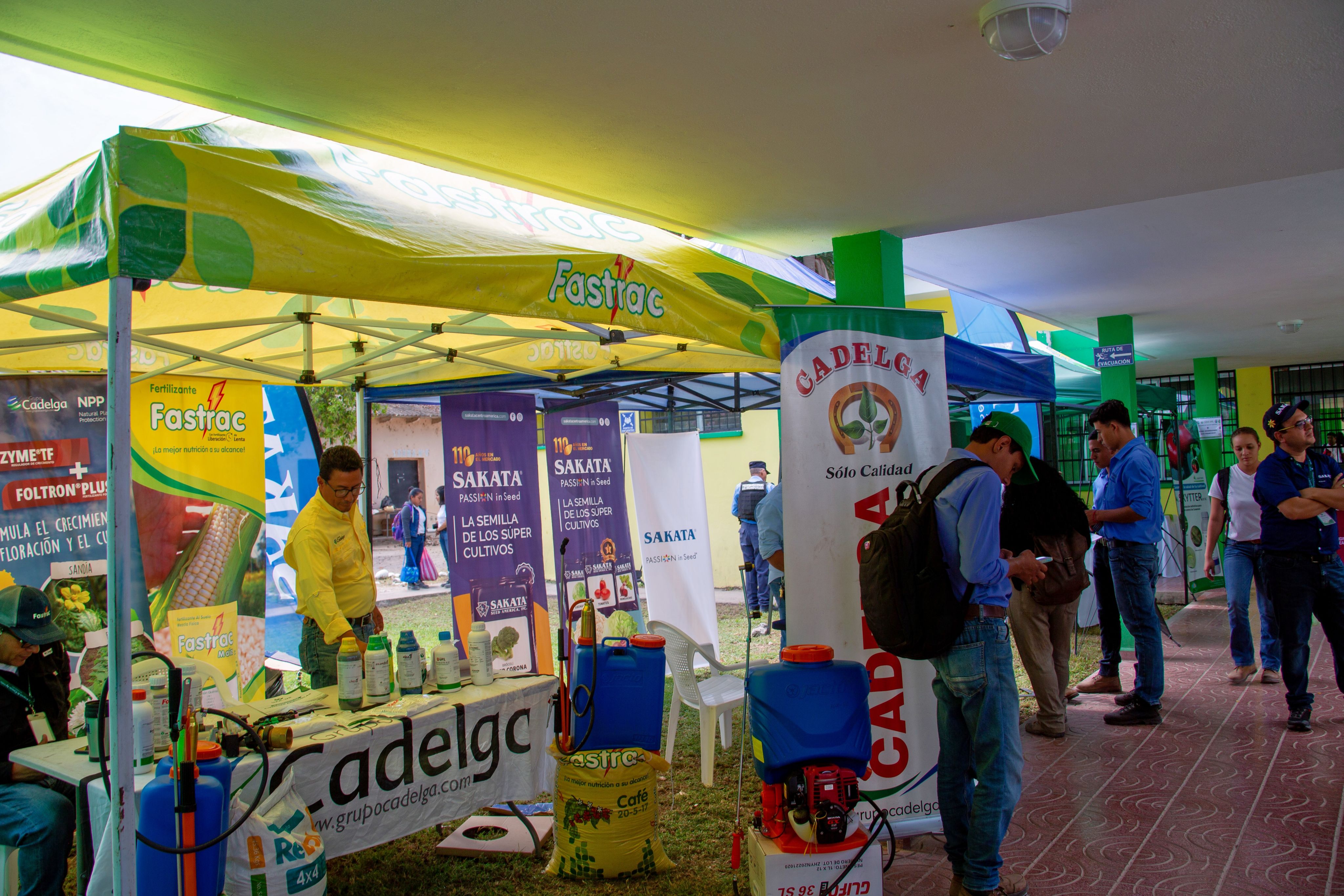 A bright yellow canopy shades vendors during the trade show exhibit at the second annual symposium in Honduras. People are walking around the various booths.