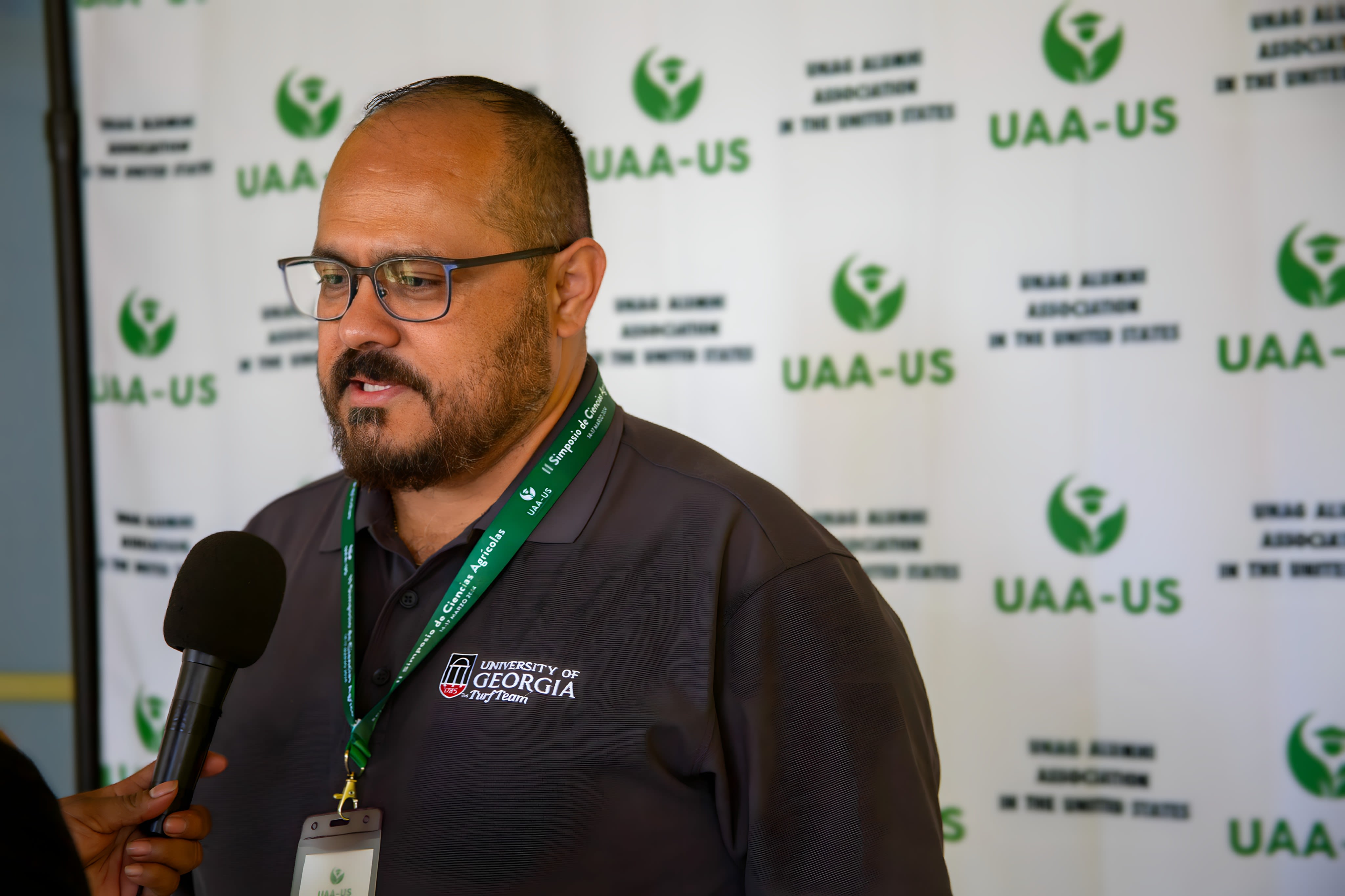 Rolando Orellana wears glasses and a black UGA Extension shirt as a microphone is held up to him during an interview. A white banner with green UAA-US logos is in the background.