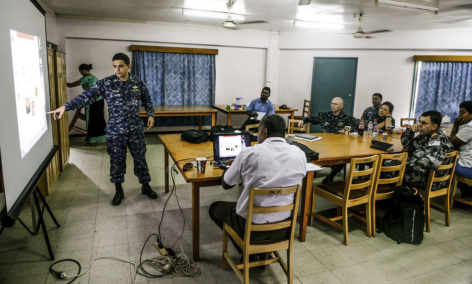 Toby Palmer stands in front of a classroom of people, some in Navy uniform, gesturing at a projector screen
