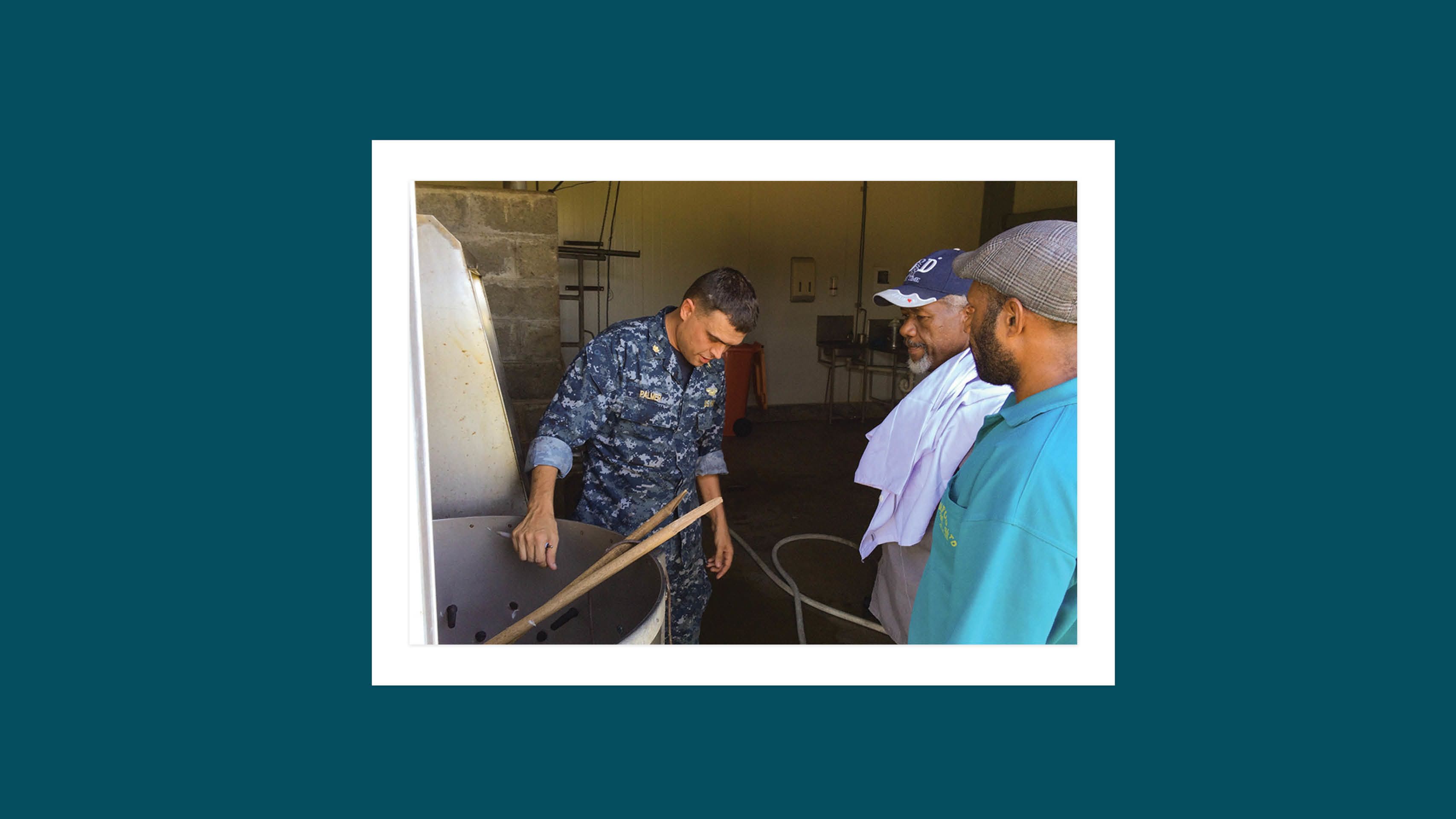 While in blue camouflage Navy uniform, Toby Palmer demonstrates a piece of equipment to two men in hats