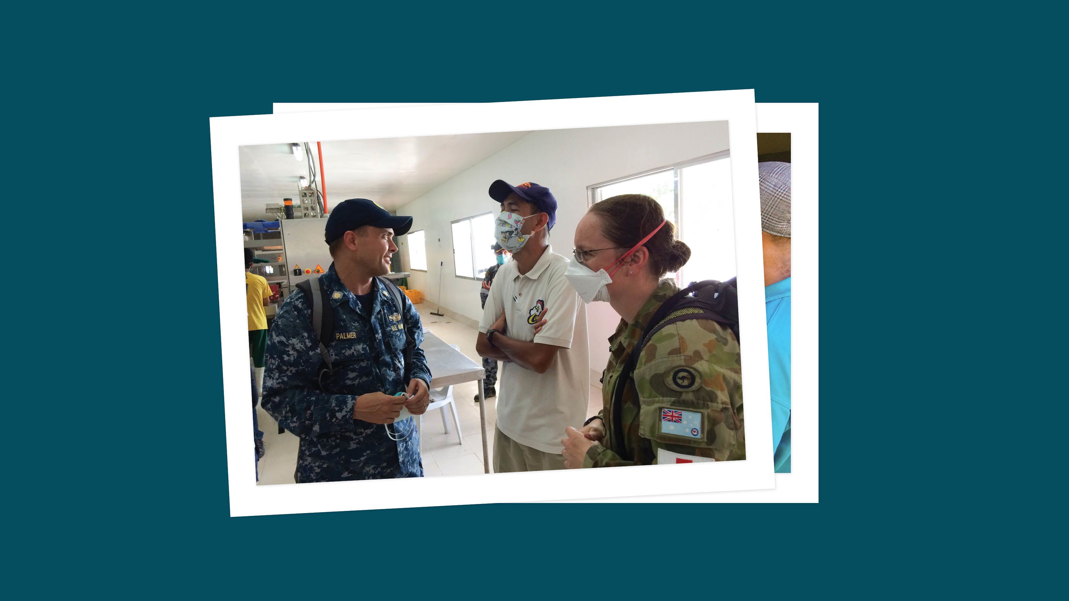 While in blue camouflage Navy uniform, Toby Palmer speaks with a man wearing a face mask and civilian clothes and a woman wearing a face mask and green camouflage military uniform. People can be seen in the background
