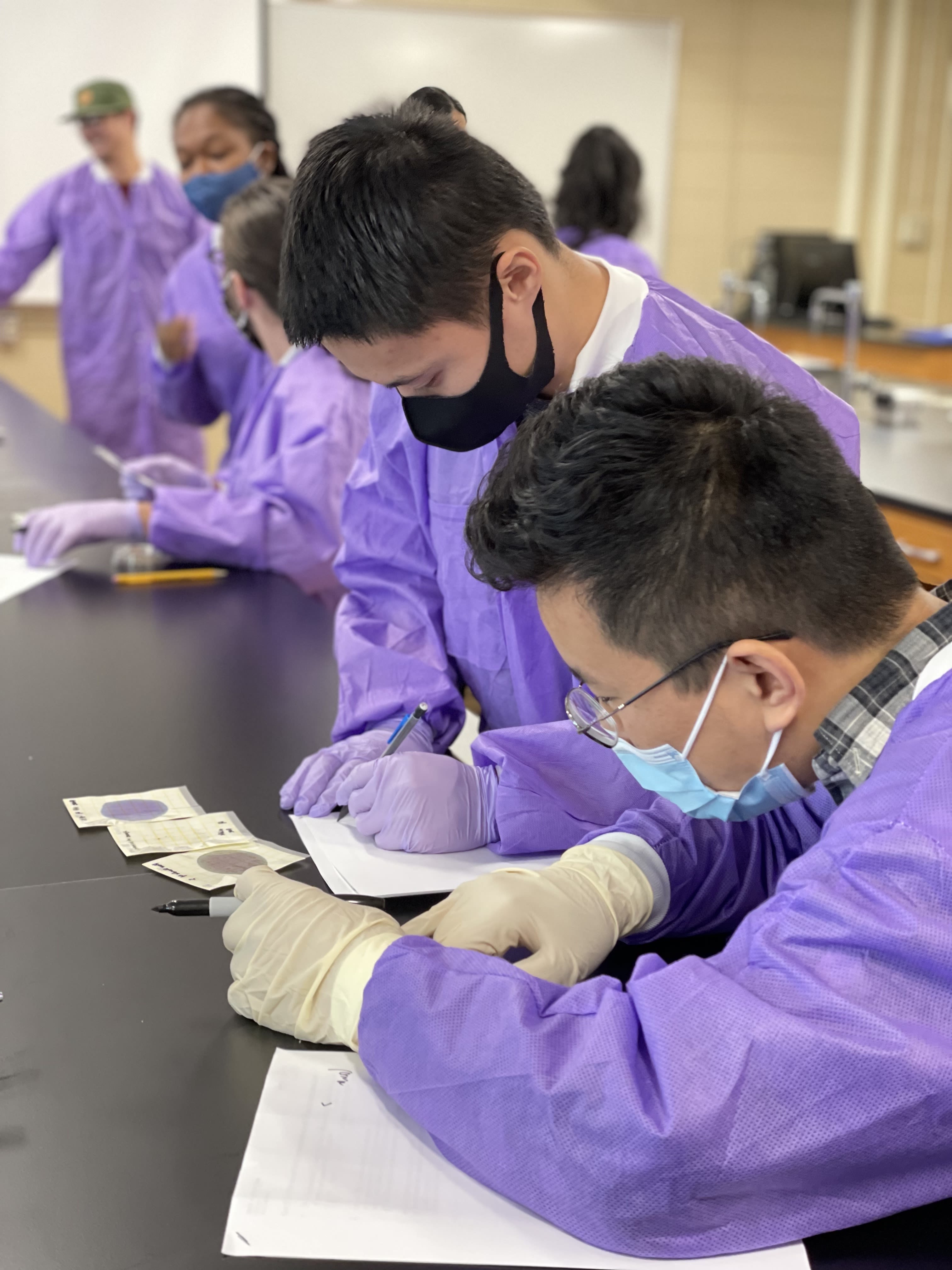 Students in purple protective outerwear and face masks make notes in a lab setting