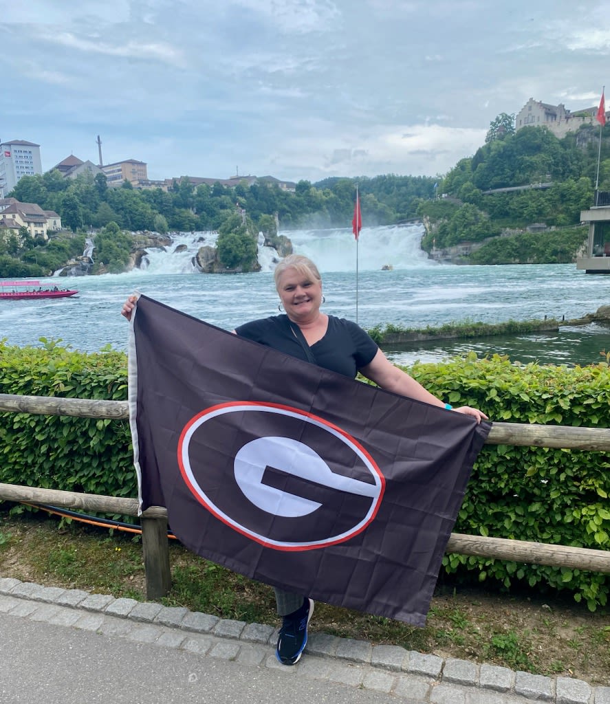 Dupree holds a large Georgia Bulldogs flag while posing in front of a fence near the banks of a river with a waterfall