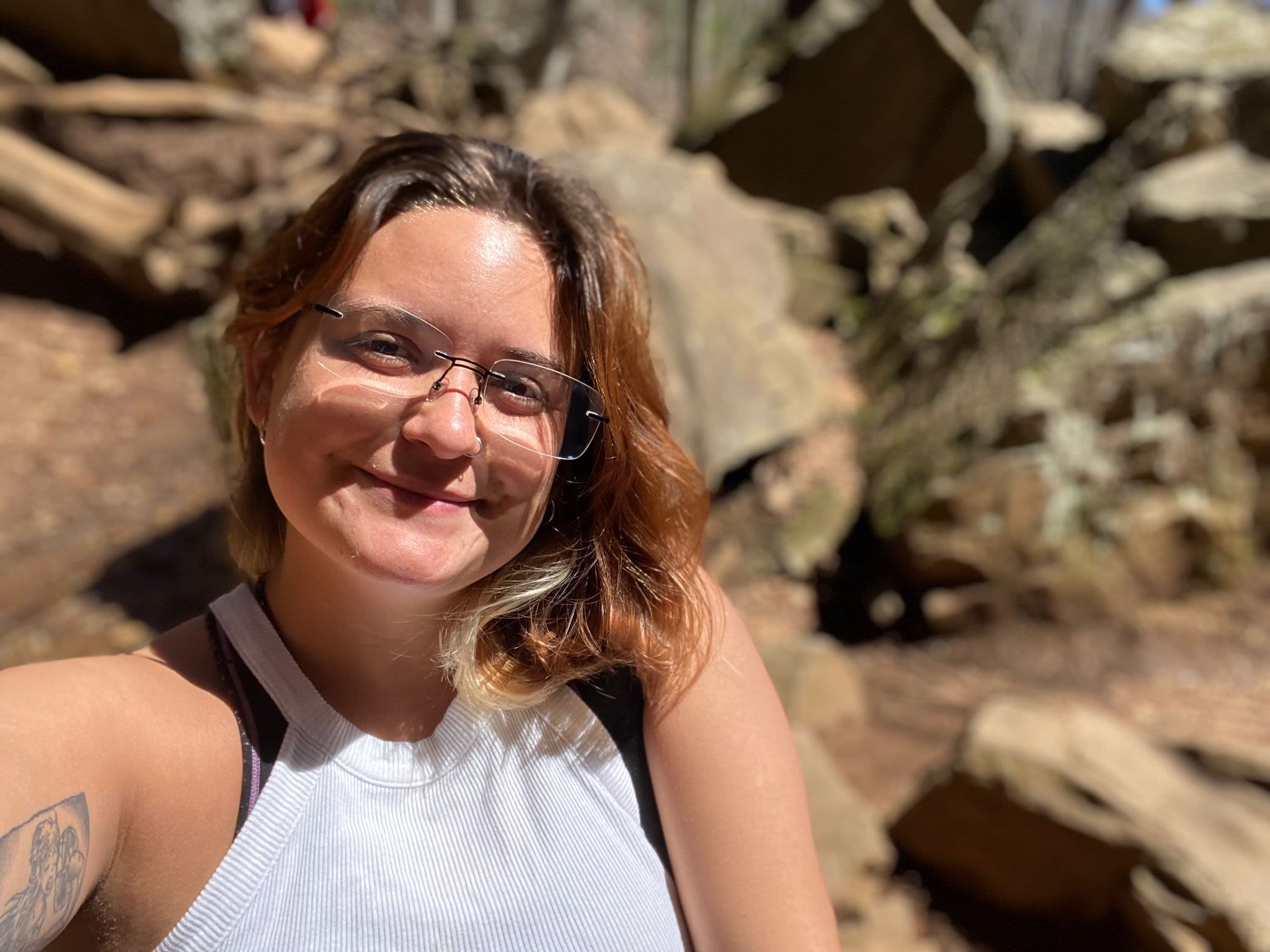 Lugo-Arroyo, wearing glasses and a white tank top, smiles while hiking on rocky terrain.