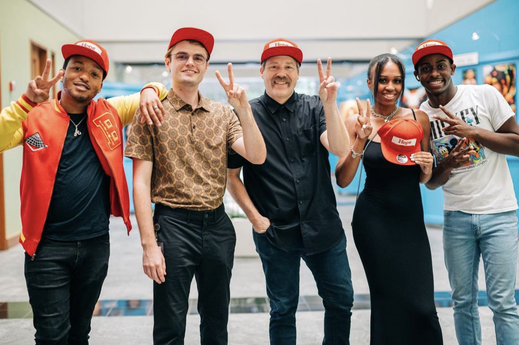 Members of the Hope Givers team, including (L-R) Brand Ambassador Nosakhere Andrews (Mr. 2-17), Program Manager Elliott Tranter, Founder and CEO Tamlin Hall, and Teen Content Team Lead Tobias Brown give their signature peace sign while wearing red Hope Givers caps and casual clothing indoors.